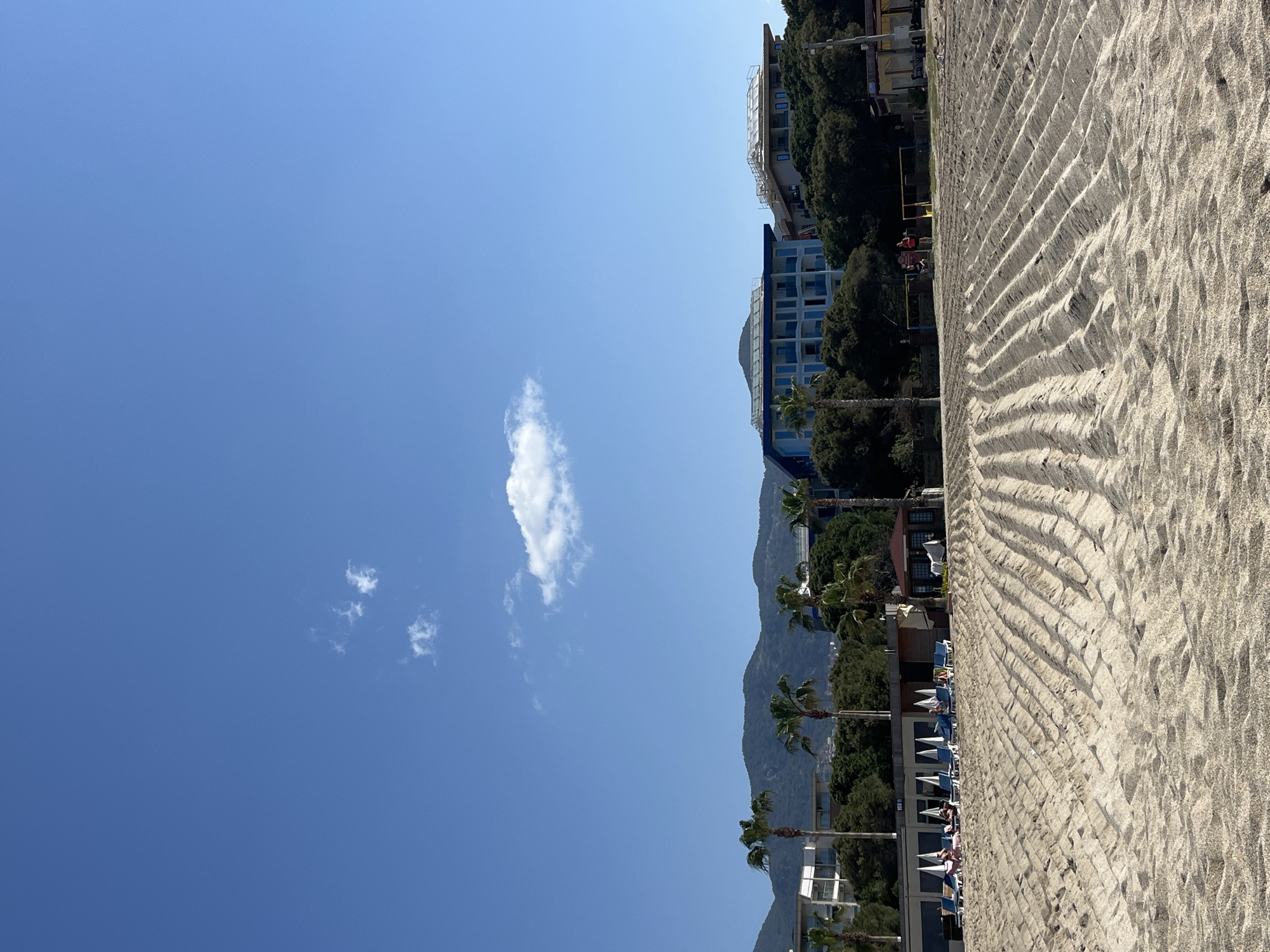 The image depicts a beach scene in Alanya, Turkey. The composition features a clear, blue sky with a small cloud on the left, transitioning to a shoreline with a sandy beach on the right. Palm trees and buildings are visible along the coastline. The beach exhibits a textured pattern in the sand. In the middle of the beach, two people are playing volleyball. Mountains are visible in the background. Deck chairs and umbrellas are set up near the shore.