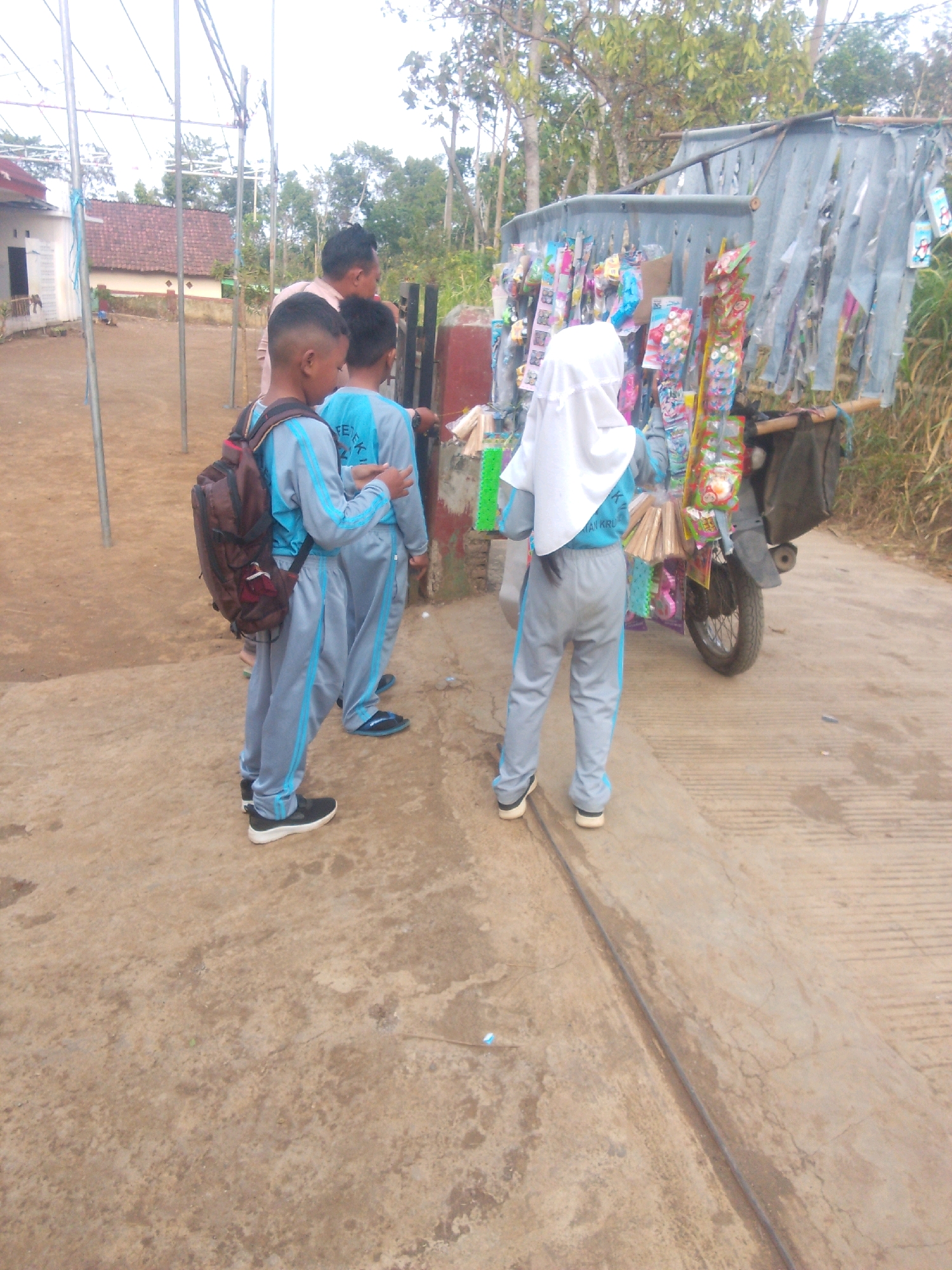 In Betek, Indonesia, an unidentified man, possibly a vendor, stands near a makeshift stall fashioned from corrugated metal and bamboo, displaying an assortment of candies and trinkets. Beside him, three young children, likely students based on their matching gray and blue uniforms, are gathered. Two boys, one carrying a brown backpack, stand to the left, facing the stall. A third child, wearing a white hijab, is on the right, facing the stall and appearing to interact with the merchandise. The environment suggests a rural or semi-rural setting with a dirt ground, sparse vegetation, and a partially visible building in the background. A motorcycle is integrated into the stall's structure, suggesting mobility.