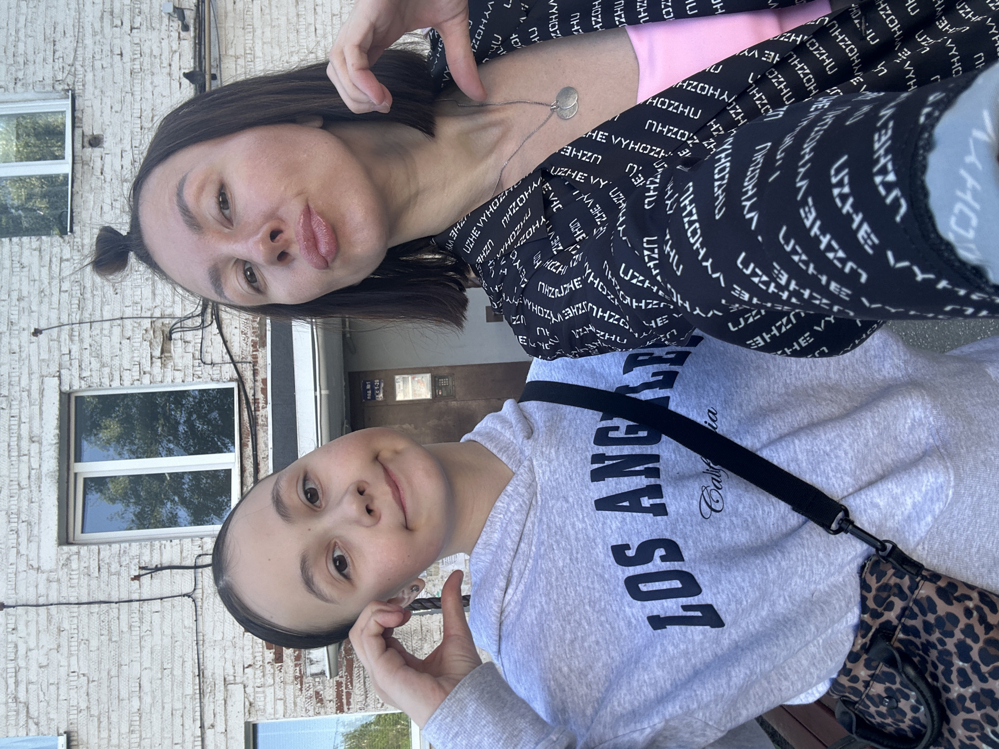 Two females pose for a selfie in front of a light-colored brick building. The younger female wears a grey "Los Angeles California" hoodie, a black shoulder strap, and a leopard print bag. The older female wears a black jacket with white text. Both make hand gestures. A window with a visible reflection is on the left. Signage in Russian is above a doorway with the number "1-20". The location is Votkinsk, Russia.