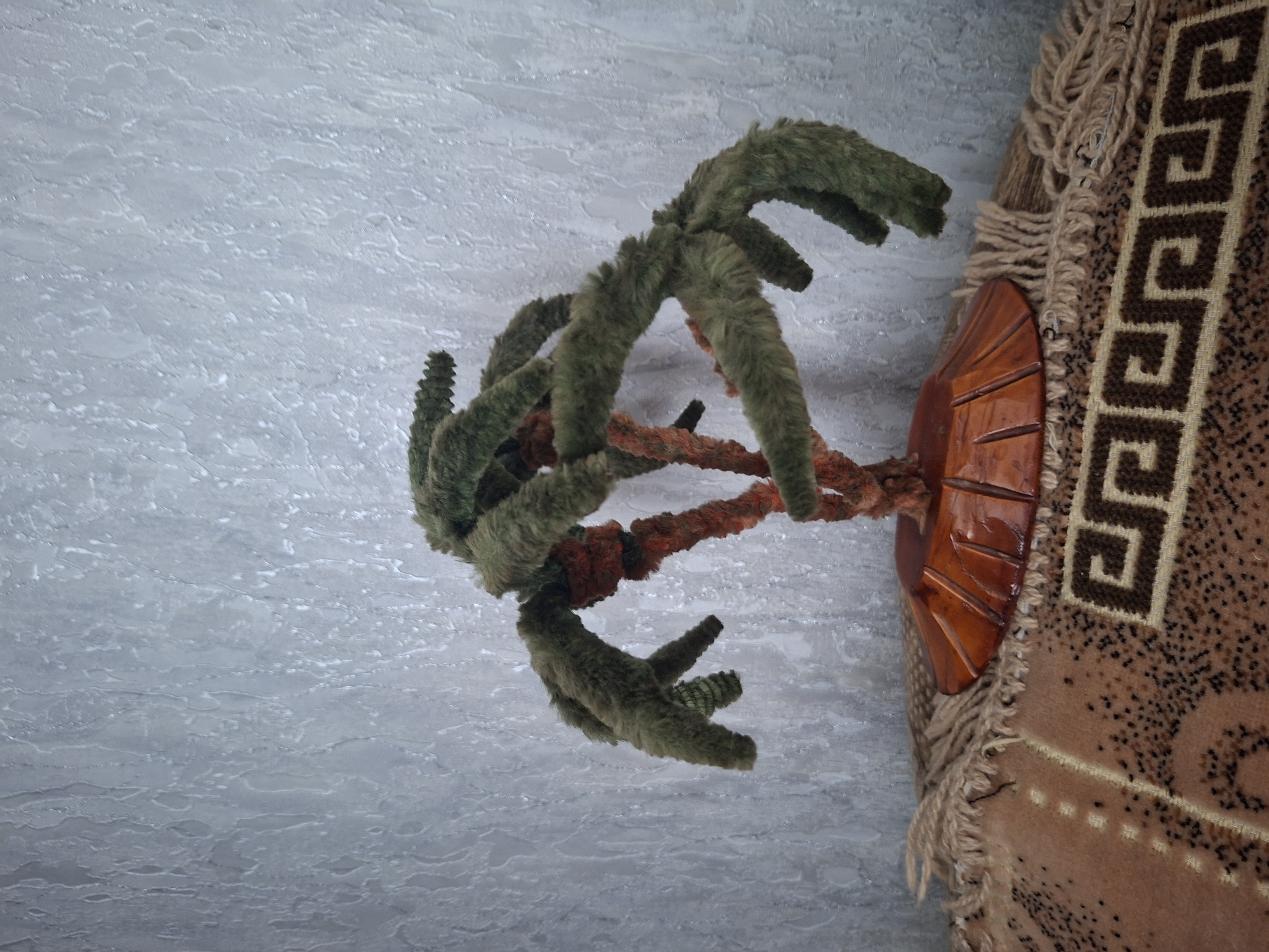The image shows a close-up, upward-facing shot of a decorative object against a textured, pale blue background. The object appears to be a faux plant with fuzzy, green, branch-like appendages that radiate outwards. Some of these appendages have a rougher, darker green texture at their tips, resembling small cones. The central structure of the plant is wrapped in a reddish-orange fuzzy material, suggesting a twisted stem or vine.
To the right of the plant, a portion of a woven tapestry or rug is visible. This textile is primarily a light brown color with a repeating geometric pattern in dark brown, resembling a Greek key motif. The edges of the tapestry are fringed with light brown yarn. Partially obscured by the plant and tapestry is a carved wooden element, possibly part of a decorative stand or base. It is orange-brown in color and has a fan-like shape with incised lines.
The background has a mottled, uneven texture with lighter and darker patches, possibly representing a wall or ceiling with an interesting finish. The lighting appears to be diffused, casting no harsh shadows. The scene is static, with no obvious activity taking place. The image does not contain any visible text. The orientation suggests the image was taken at an angle, possibly looking up at the object. The overall impression is of an indoor decorative arrangement, possibly with a rustic or folk art aesthetic, given the woven tapestry and carved wood.