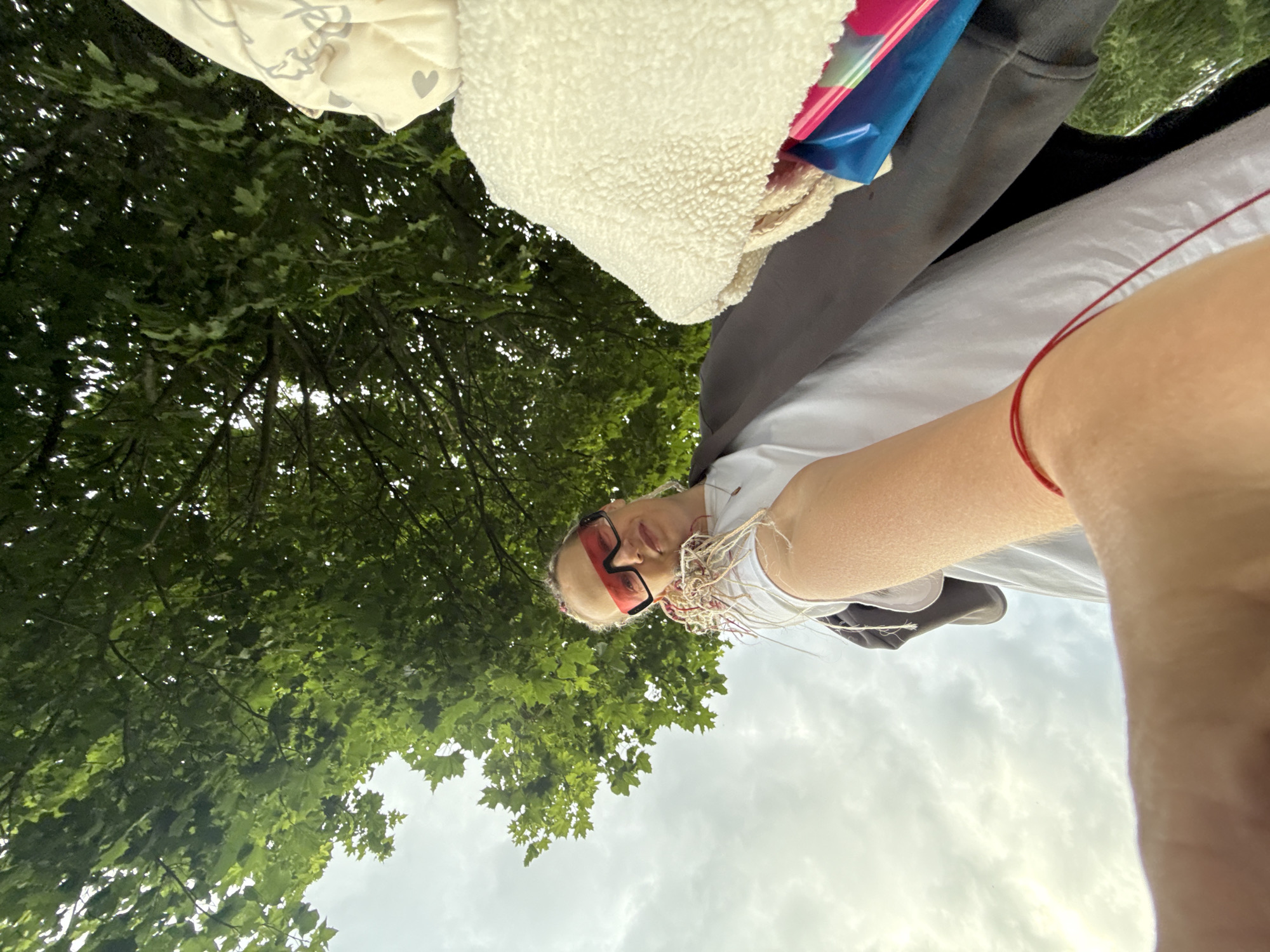An individual is captured from a low-angle perspective, looking upwards towards a canopy of green trees against a cloudy sky. The person, who appears to be female, is wearing large, bright red sunglasses and a white t-shirt with a dark jacket over it. Their hair is styled in braids with colorful strands. A fluffy white garment and a blue and pink object are visible to the right of the frame. The person's arm is extended, holding what appears to be a small bunch of sparkly decorations. A thin red string is visible wrapped around their wrist. The scene suggests an outdoor setting, possibly a park or a street with trees, in Moscow, Russia, as indicated by the context provided.