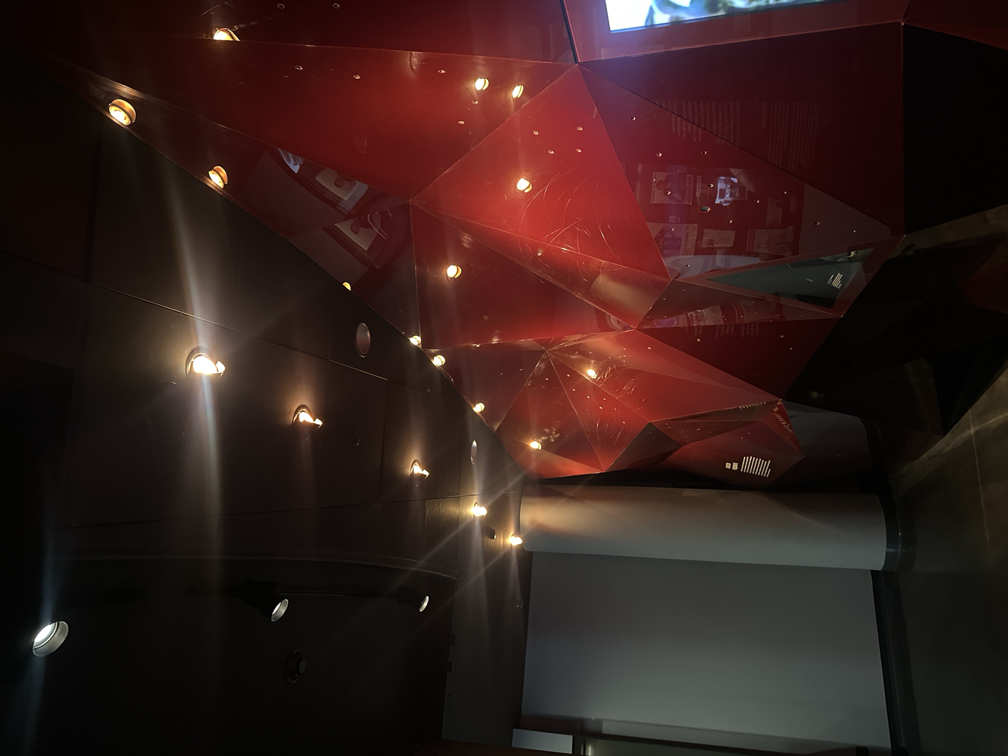 The image depicts the interior of a room, potentially a museum or exhibition space, located in Yekaterinburg, Russia. The ceiling features a complex, geometric design constructed from red, reflective panels. Small, recessed lights are integrated into both the ceiling and the adjacent wall, casting beams of light downwards. A pull-down projection screen is visible on the right side of the frame. The walls appear to be dark in color, creating a contrast with the brighter elements of the ceiling and the screen. No people are visible in the scene. The overall impression is one of modern, architectural design.