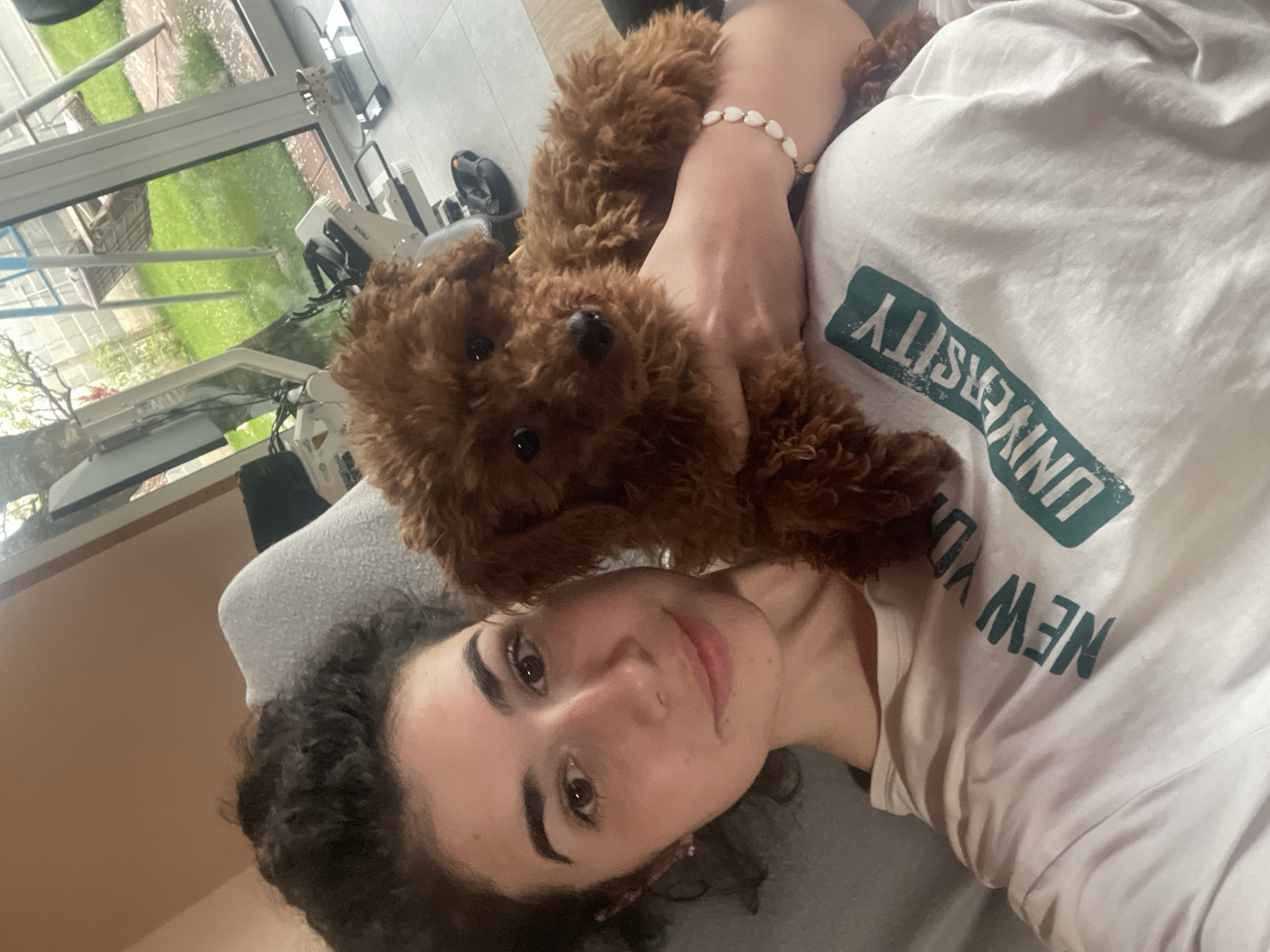 A close-up selfie-style image captures a person and a small, fluffy brown dog resting together indoors in Sofia, Bulgaria.
The main subjects are a young person with dark curly hair, prominent eyebrows, and a slight smile, and a small, brown, curly-haired dog, likely a poodle or similar breed, with dark eyes and nose. The person is lying down, wearing a white t-shirt with the text "NEW YORK UNIVERSITY" printed in green, visible upside down due to the selfie angle. A bracelet made of small, heart-shaped, light-colored beads is visible on the person's wrist.
The dog is positioned on the person's chest or shoulder, looking directly at the camera, while the person holds their hand close to the dog. The overall activity appears to be a relaxed, affectionate moment, possibly a cuddle session or playtime captured in a selfie.
In the background, a large window offers a view of an outdoor area with vibrant green grass, indicating a natural setting like a garden or lawn. Some light-colored specks are visible on the grass, possibly pollen or blossoms. Several pieces of indoor equipment, including what looks like a floor lamp or stand and other devices, are partially visible near the window. The natural light filtering through the window suggests it is daytime. The emotions conveyed are generally calm and happy.