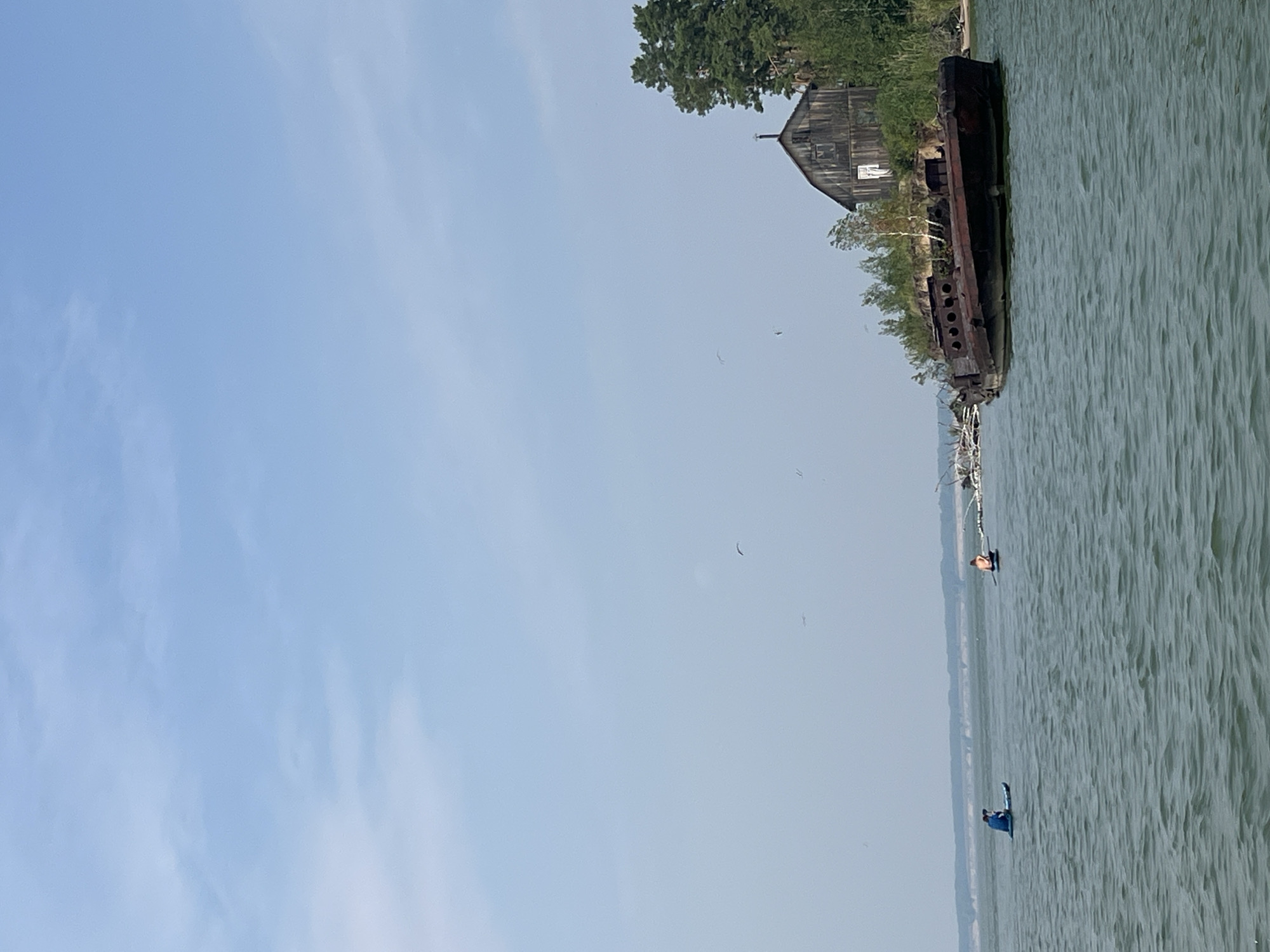 The image shows a body of water with two people on paddleboards. In the background, there is a shipwreck and a wooden structure resembling a barn or house on a small, vegetated island. The sky is clear with some clouds, and there are birds in the distance. The water is choppy, and the lighting suggests it is daytime. The scene appears to be in a natural, perhaps remote, setting, and is not indicative of the city of Borovoe, Russia, which is known for its mountainous terrain and clear lakes.