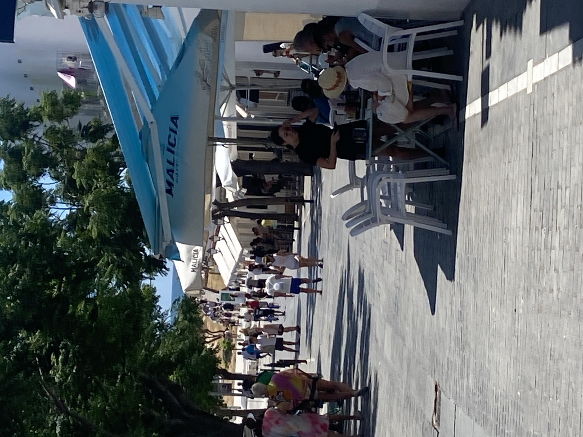 The image captures a sunny outdoor scene in Conil de la Frontera, Spain, likely on a promenade or street. A walkway paved with light-colored stones dominates the foreground, with shadows cast by trees and structures. Several people are walking and standing. On the right, a white cafe awning with blue stripes is visible, featuring the word "MALICIA" in blue letters. Beneath this awning, a woman in a black top and shorts stands near a table with customers. She appears to be a server, holding a dark object, possibly a phone or order pad. A woman in a white outfit and a straw hat sits at the table, facing away from the camera.
Further into the scene, a diverse group of people stroll along the walkway. Some are casually dressed, suggesting a vacation atmosphere. Trees line the left side of the walkway, their dense foliage providing shade. In the background, buildings with white facades are visible, hinting at shops or residences. The ocean is faintly visible in the distance beyond the trees and buildings. The overall impression is one of a lively, pleasant day in a coastal Spanish town.