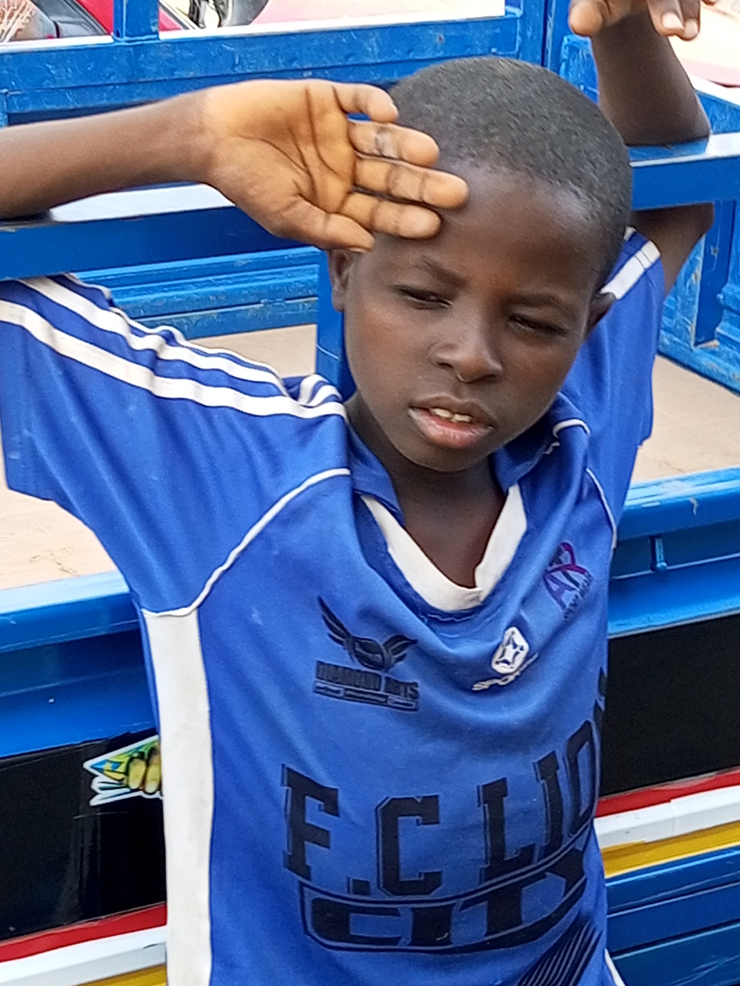 A young boy, appearing to be of African descent, stands leaning against a blue structure. He is wearing a blue shirt with white stripes on the sleeves and a white collar. His right arm is raised, with his hand resting on his forehead, partially obscuring his eyes. The background is a blurred view of a red vehicle and what appears to be the side of a truck with colorful stripes. The setting seems to be outdoors. The boy's gaze is directed downwards and to his left. The lighting suggests it is daytime. There is no visible indication of the location being in Arawa, Nigeria.