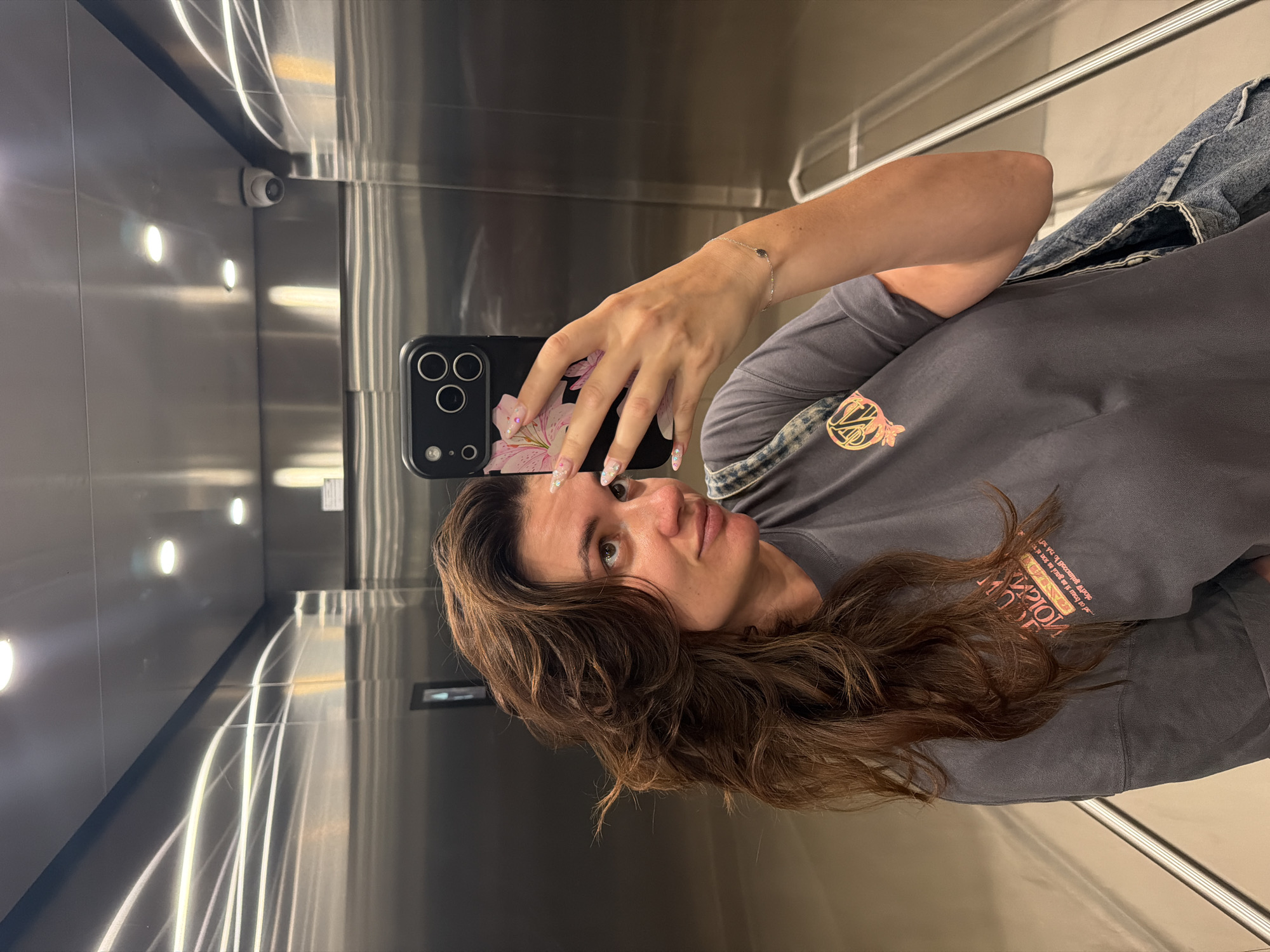 A woman takes a selfie in a stainless-steel elevator. She has long, dark brown hair that is styled with loose waves and is looking to her right. She is wearing a grey t-shirt and has a denim jacket slung over her shoulder. Her phone, with a floral-patterned case, is held up to take the photo. The elevator interior is highly reflective, showing the lights and the camera on the ceiling. The image was captured in the city of Belgrade, Serbia.