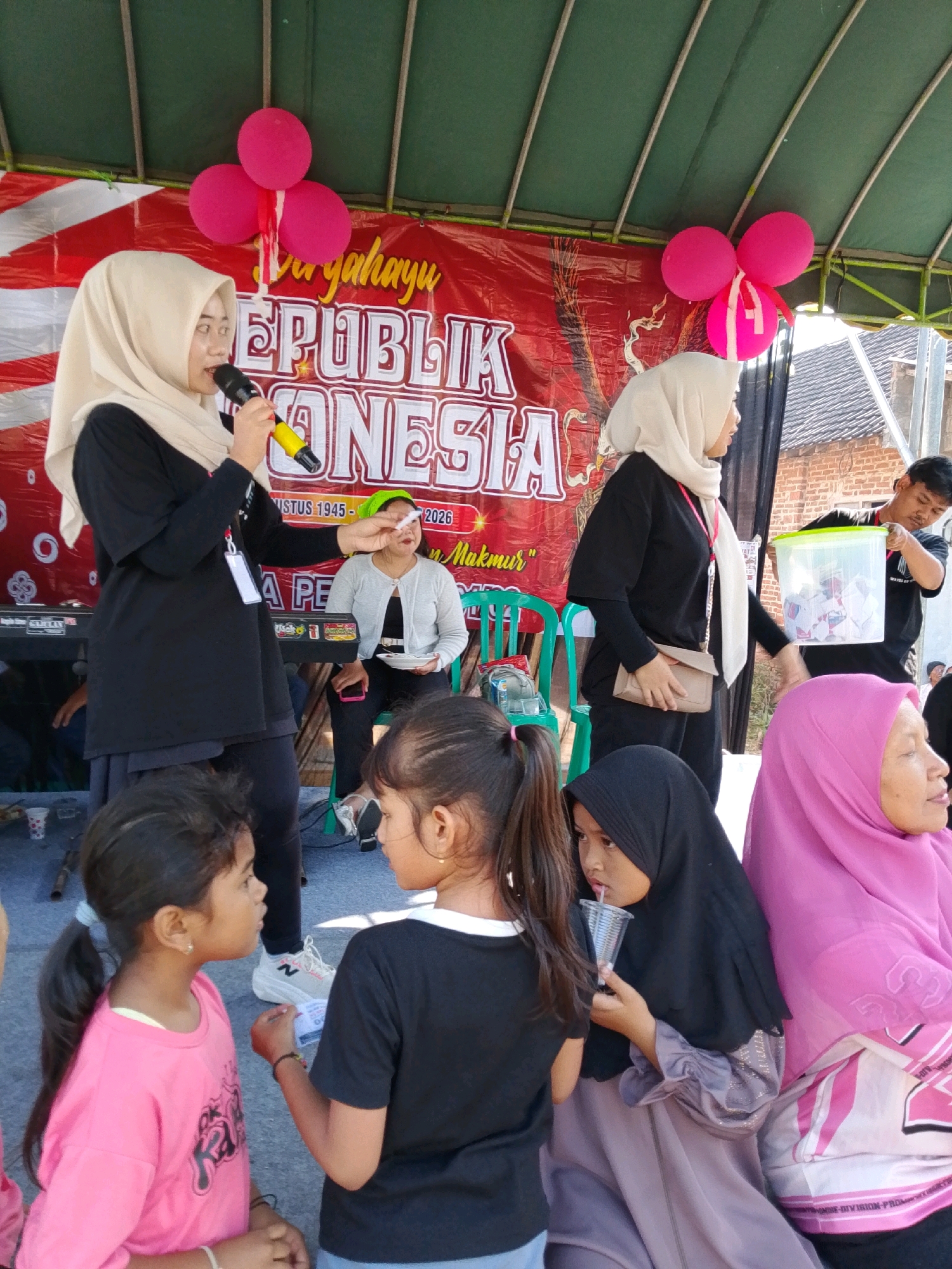 In Banyakan, Indonesia, on a sunny day, a group of people is gathered under a green canopy. In the foreground, two young girls are interacting. The girl on the left wears a bright pink shirt with graphics and has her hair in a ponytail. The girl on the right wears a black shirt and is talking to her. Behind them, several women and a man are present. One woman, wearing a light beige hijab and a black outfit, is speaking into a microphone. Another woman, wearing a grey cardigan, sits on a chair holding a white plate. To the right, a man in a black shirt is holding a clear plastic container filled with slips of paper. A woman in a pink hijab is seated to the right of the container. A banner with red and white stripes and text that reads "Jaya Republik Indonesia" is visible behind the performers. Pink balloons are attached to the canopy. The environment appears to be outdoors, possibly a community event or celebration.