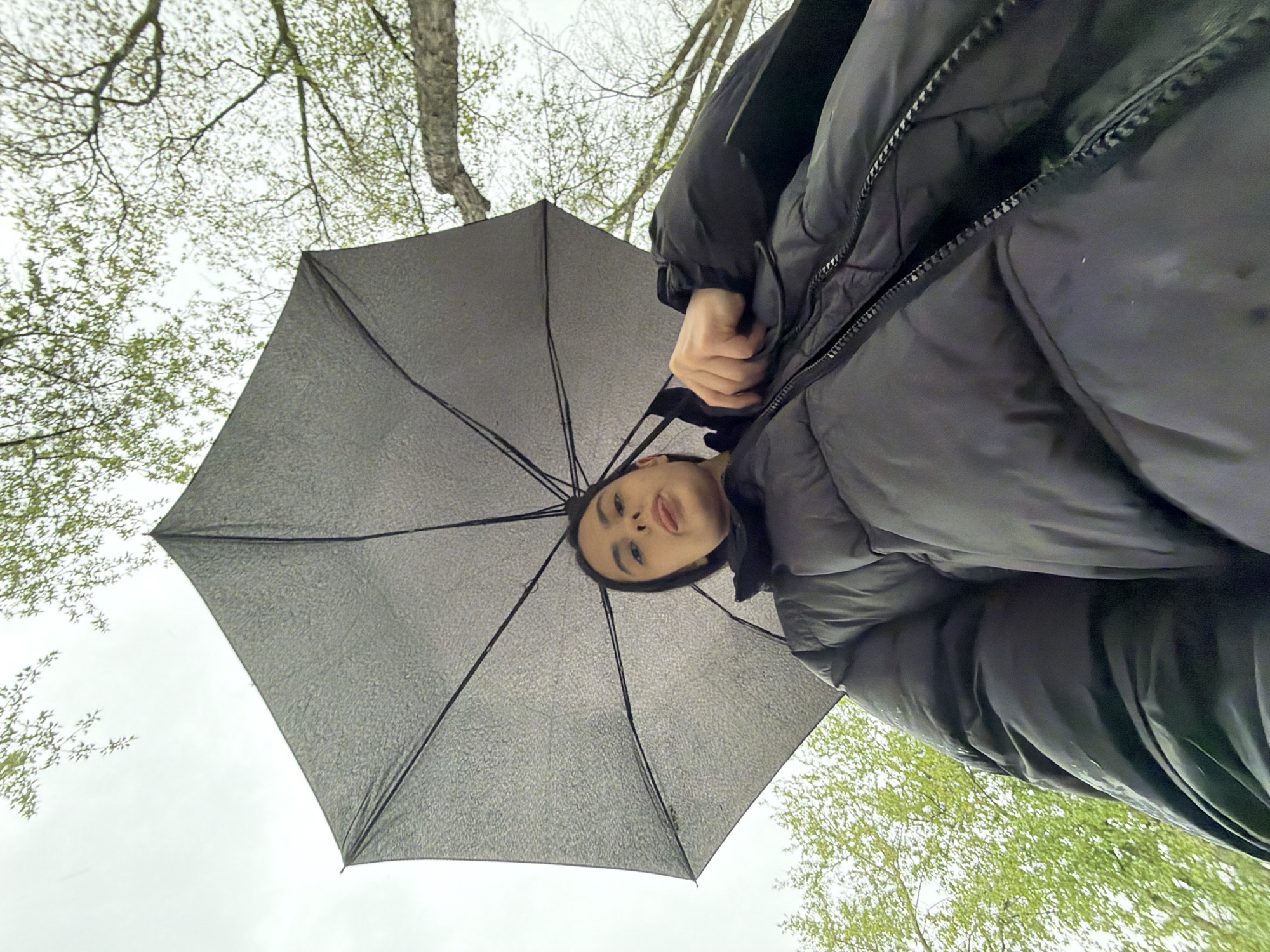 A low-angle shot shows a woman holding a gray umbrella overhead, presumably on a cloudy day. The woman has dark hair and is wearing a dark puffer jacket. The background includes tree branches with budding green leaves against an overcast sky. This photo was taken in Chaikovsky, Russia.