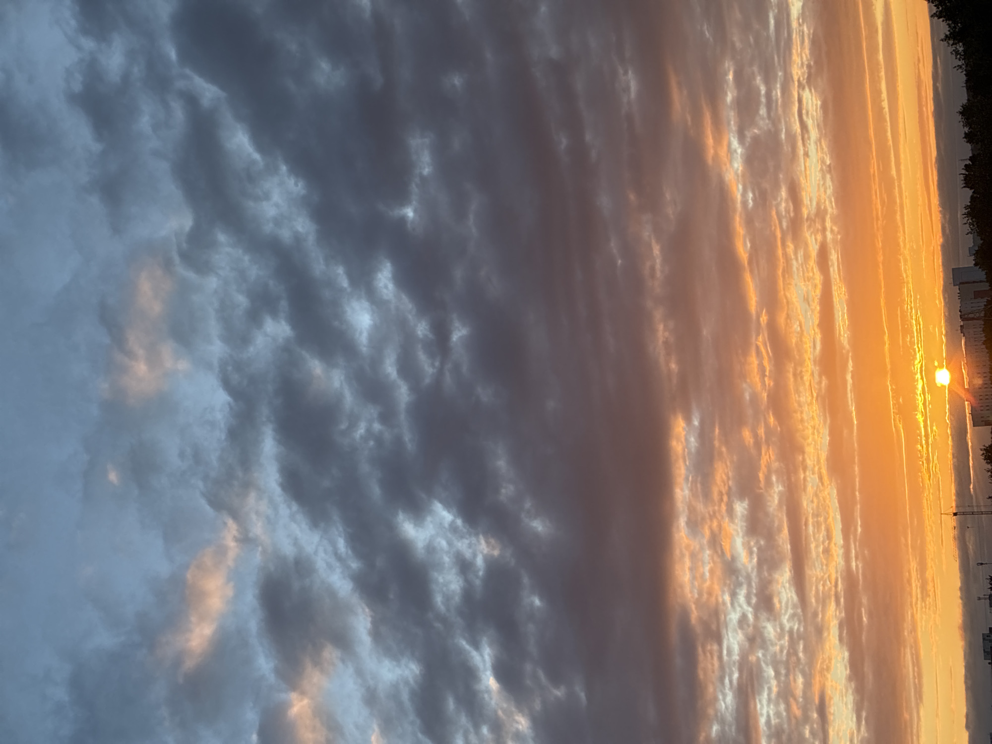 The image depicts a dramatic sky over an urban landscape. The upper portion of the sky is a pale blue with wisps of dark gray clouds. Moving downwards, the clouds become denser and take on hues of pink and orange as they are illuminated by the setting sun. The sun itself is a bright, blinding white orb peeking from behind the clouds on the right side of the frame, casting a vibrant orange glow across the horizon. Below the sky, a dark silhouette of trees and buildings is visible, with a few taller structures, including what appears to be a construction crane, standing out against the colorful sky. The overall impression is of a city at sunset, with the sky being the dominant and most visually striking element.