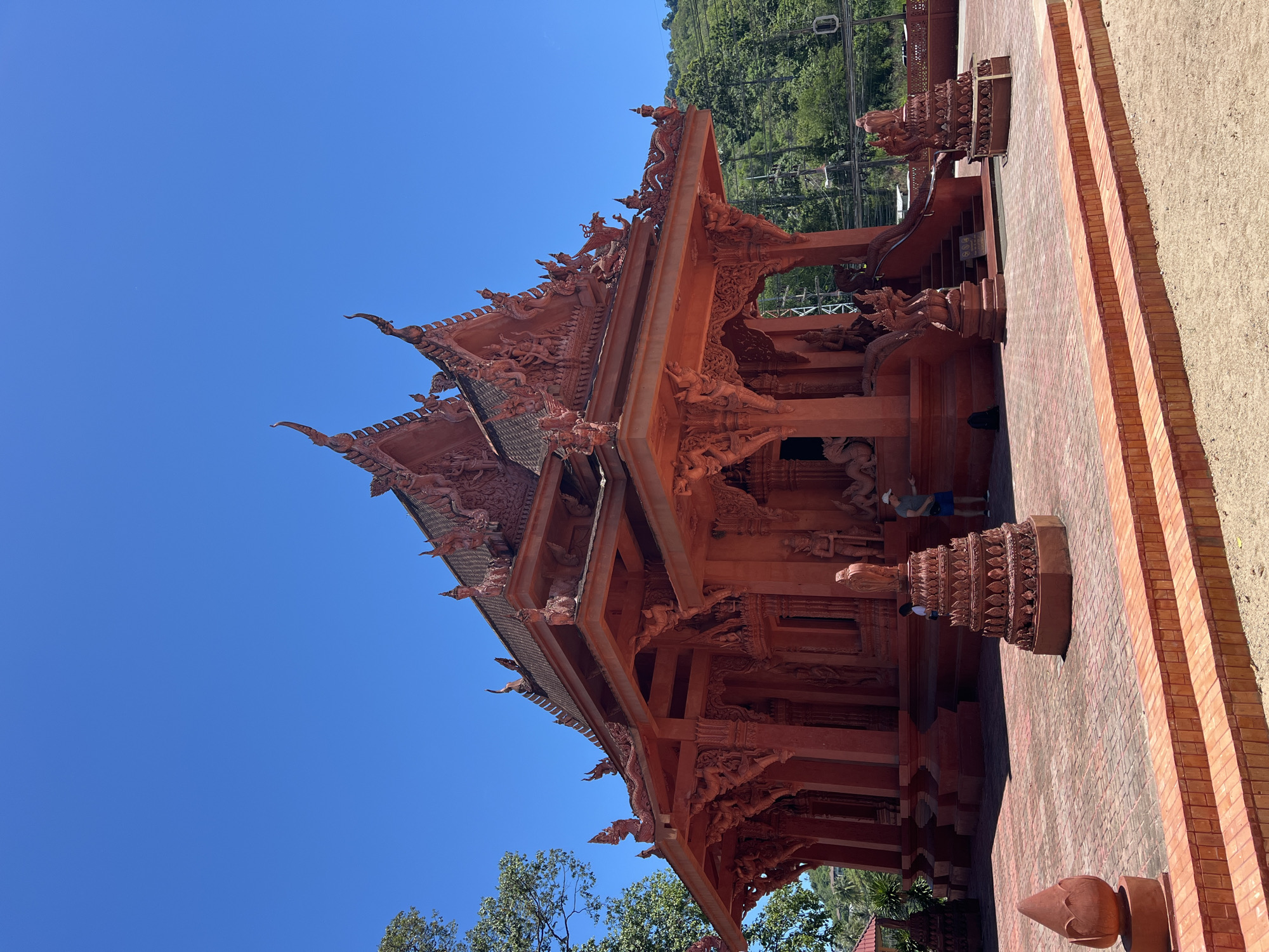An ornate, bright red Buddhist temple stands under a clear blue sky in Baan Hua Thanon, Thailand. The temple features intricate carvings and decorative rooflines with tiered spires. In front of the temple, a paved courtyard is decorated with red brick patterns and several ornamental red statues. A person in a white shirt and blue shorts stands on the steps to the temple, and another person in a white shirt is visible further back. To the left of the temple, a lush green tree adds a natural element to the scene, and a hill covered in dense vegetation rises behind the temple on the right. The overall impression is one of vibrant color and detailed craftsmanship in a warm, sunny environment.