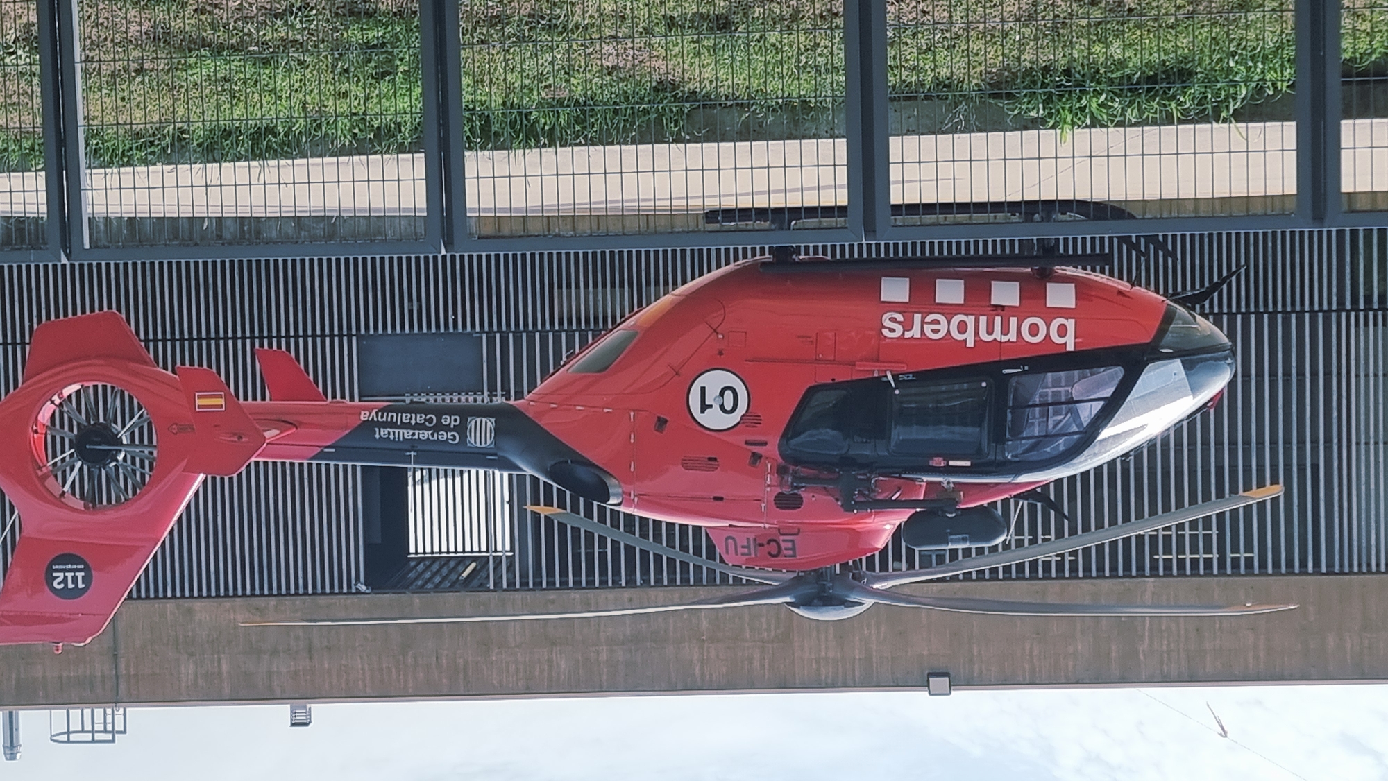 The image shows a red and black helicopter parked in front of a modern building with vertical metallic siding. The helicopter has "bombers" written in white on its side, along with a round marking with "C1" inside. It also displays "EC-IFU" near the rotor and "Generalitat de Catalunya" on the tail boom. A "112 emergencies" label is visible on the tail fin. A Spanish flag is also present on the tail. The helicopter is behind a wire fence, with green grass and plants in the foreground. The sky is partly cloudy. This scene is likely at an emergency services base or airport in the city of Olot, Spain, given the markings on the helicopter. There are no people visible in the image.