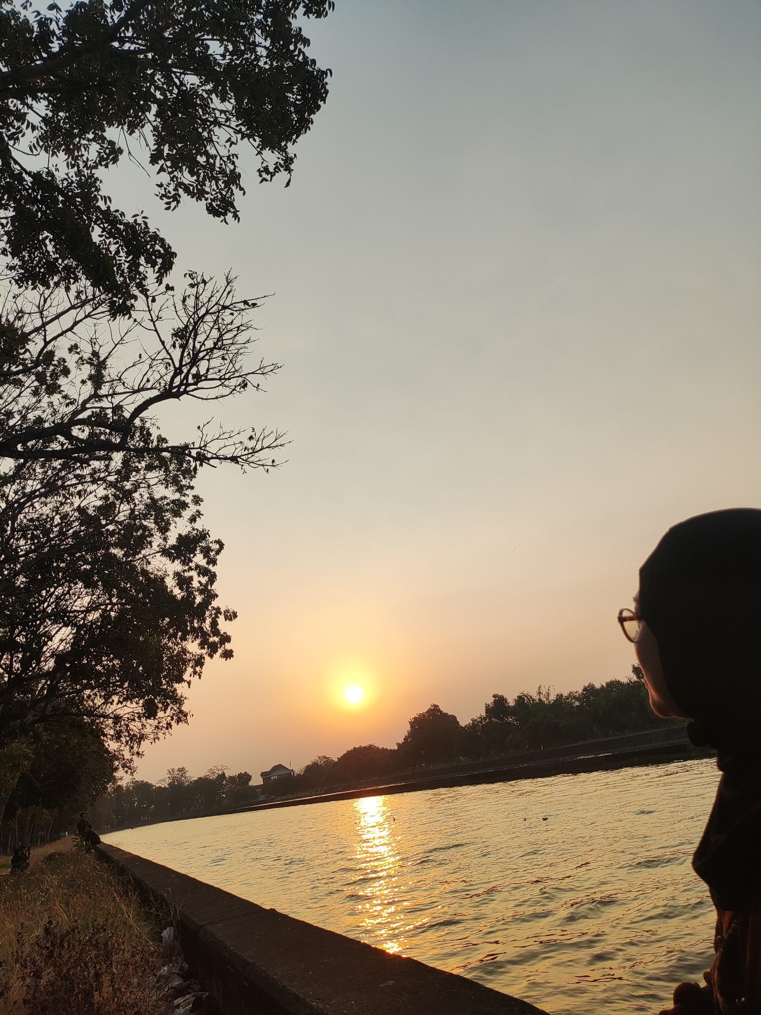A person wearing a black hijab and glasses looks out over a body of water during sunset. The sun, a bright orb, is positioned in the left-center of the frame, casting a golden glow on the water's surface, creating a shimmering effect. The water appears calm with gentle ripples. Along the bank of the water is a concrete barrier, with dry grass and some discarded trash visible on the left side of the barrier. Beyond the water, a line of trees forms the horizon, with some buildings and structures visible amongst them. In the upper left corner of the image, the dark silhouette of tree branches with leaves extends into the pale sky, which transitions from a lighter hue at the top to a warm, orangish tone near the horizon. The scene is likely a public park or recreational area within the city of Panongan, Indonesia, given the presence of the water body and the natural elements.