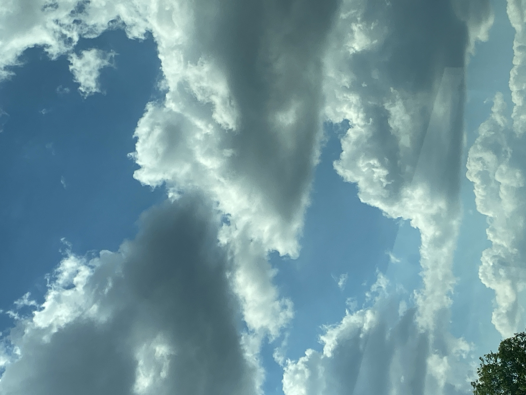 A cloudy sky is visible, with a patch of blue sky peeking through. The clouds are a mix of white and gray, with some appearing more dense than others. In the bottom left corner, the tops of some green trees are visible, suggesting an outdoor scene. The image is taken from a low angle, looking upwards towards the sky. The location is identified as Dobkowice, Poland. There are no people or specific objects clearly discernible within the scene, beyond the natural elements of sky and trees. The image captures a moment of a partly cloudy day.