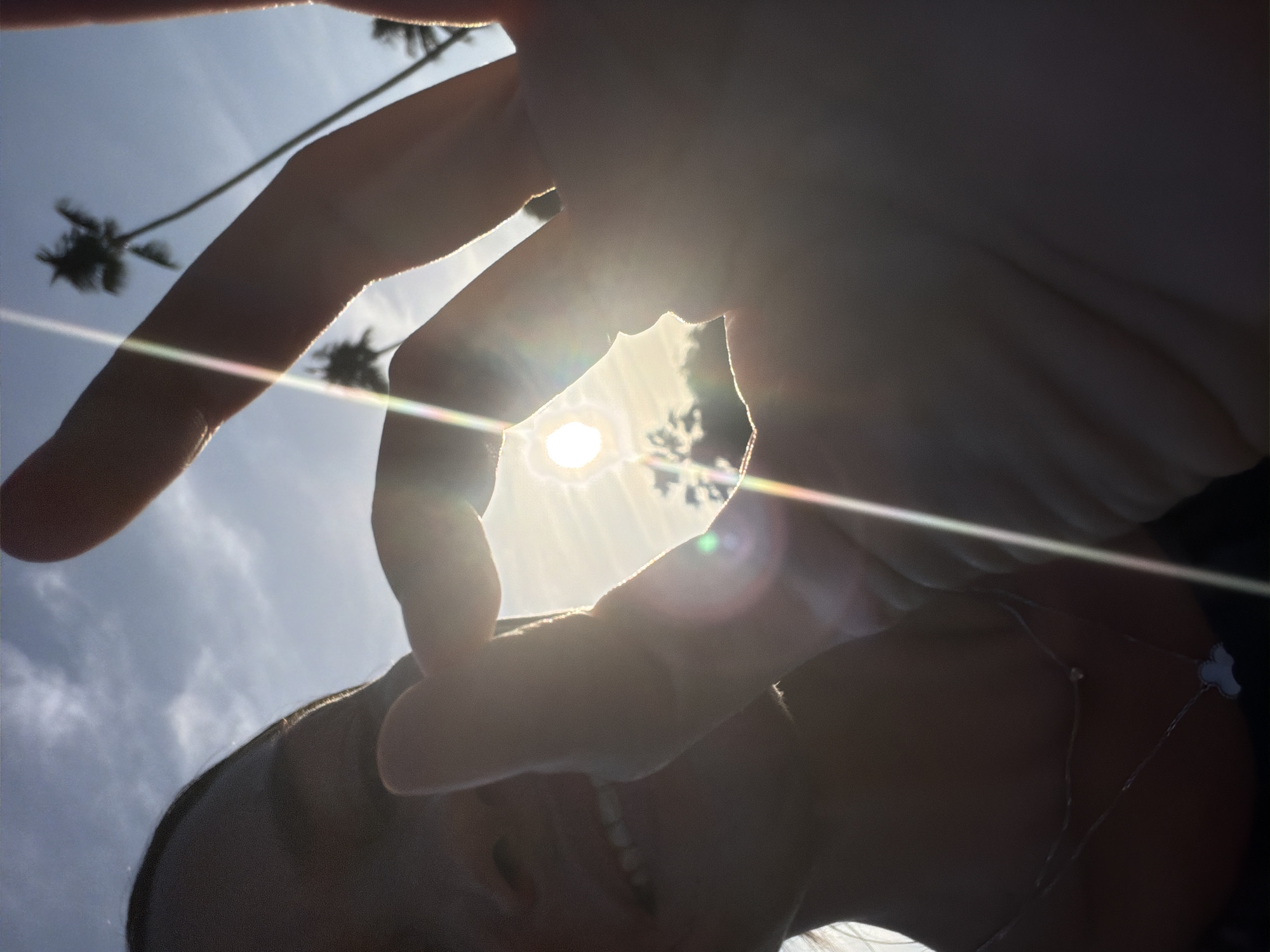 A low-angle, silhouetted selfie captures a person holding their fingers to form a circle, through which the sun is visible. The sun is bright and creates lens flare, with rainbow-like streaks of light extending from it. Palm trees are silhouetted against the bright sky in the background, suggesting a tropical location. The person's face is mostly in shadow, but their smile is visible. They are wearing a necklace. The overall lighting and palm trees suggest the scene is set in a warm, sunny environment, possibly Ko Samui District, Thailand, during daytime.