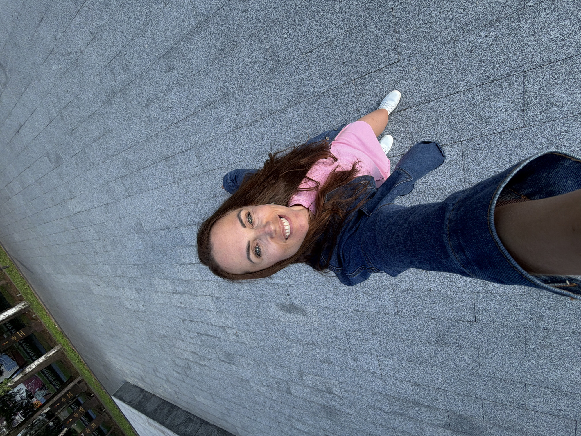 In Birobidzhan, Russia, a woman with long brown hair, wearing a denim jacket over a pink dress and white sneakers, takes a selfie while standing on a paved area. The ground is made of large, grey rectangular tiles arranged in a grid pattern. To the left of the woman, a raised concrete structure is visible. In the background, trees and what appear to be grave markers or monuments are present. The image is taken from a high angle, looking down at the woman and the surrounding area.