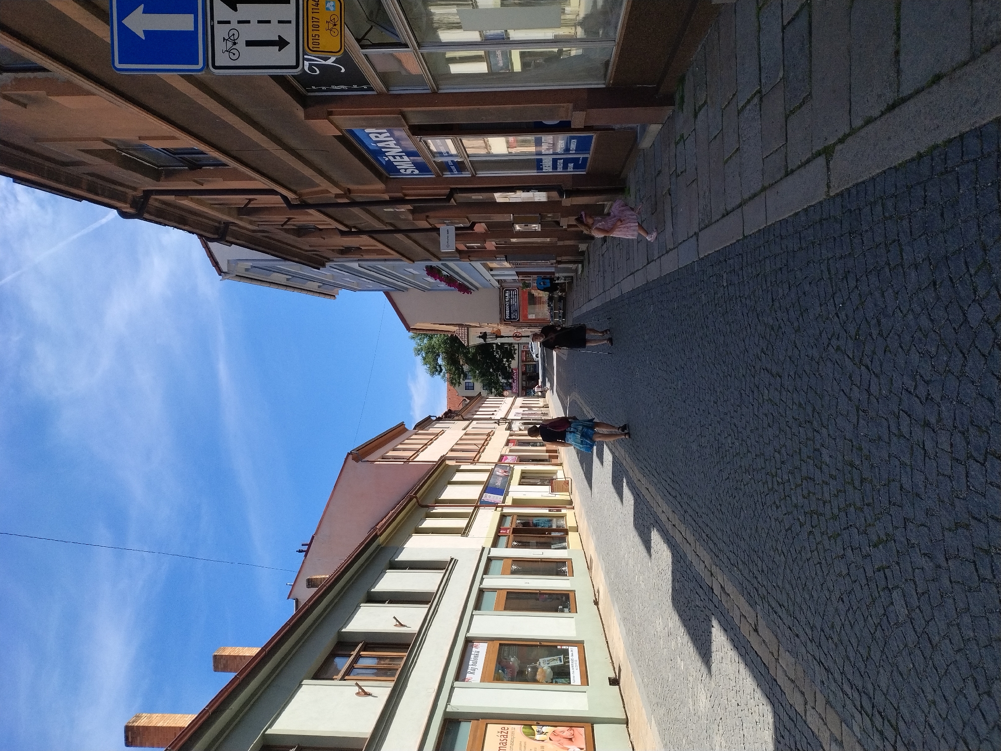 The image depicts a street in Písek, Czechia, featuring traditional European architecture. The street is paved with cobblestones and lined with buildings on both sides, displaying a mix of historical facades and modern storefronts. On the left, a light green building has several windows and what appear to be shop displays. Further down, a pastel-colored building also features shops. On the right, a darker brick building houses a currency exchange shop named "SMĚNARNI". Road signs are visible on the right, including a directional arrow and a bicycle symbol. Several people are present: a woman in a blue skirt walks away from the camera on the left side of the street, while another woman and a young girl in a pink dress are walking on the sidewalk on the right. The sky is clear with some wispy clouds, suggesting a sunny day. The perspective of the image looks down a straight street, giving a sense of depth and leading the eye towards the distant buildings.
