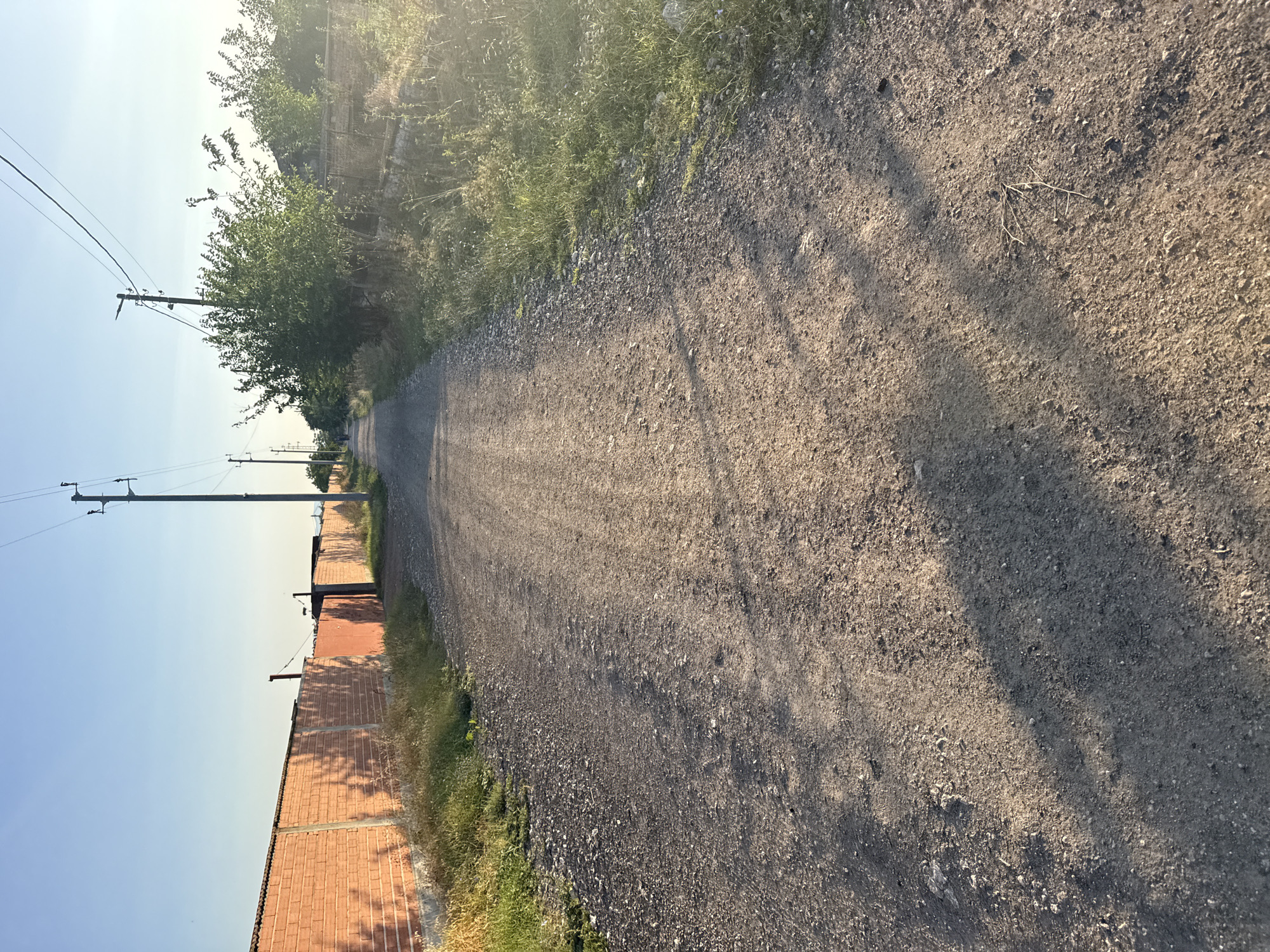 The image depicts a dirt road in Shishmanți, Bulgaria, with a clear blue sky overhead. On the left side of the road is a long, orange brick wall. On the right side, there are overgrown weeds and trees. Several utility poles with power lines are visible along the road. The road itself is unpaved and appears to be a residential access road, with tire tracks visible on its surface. Shadows from the trees cast dappled patterns on the road, indicating it is late afternoon or early morning. In the distance, a person is walking away from the camera on the road. The overall scene is a rural or semi-rural setting.