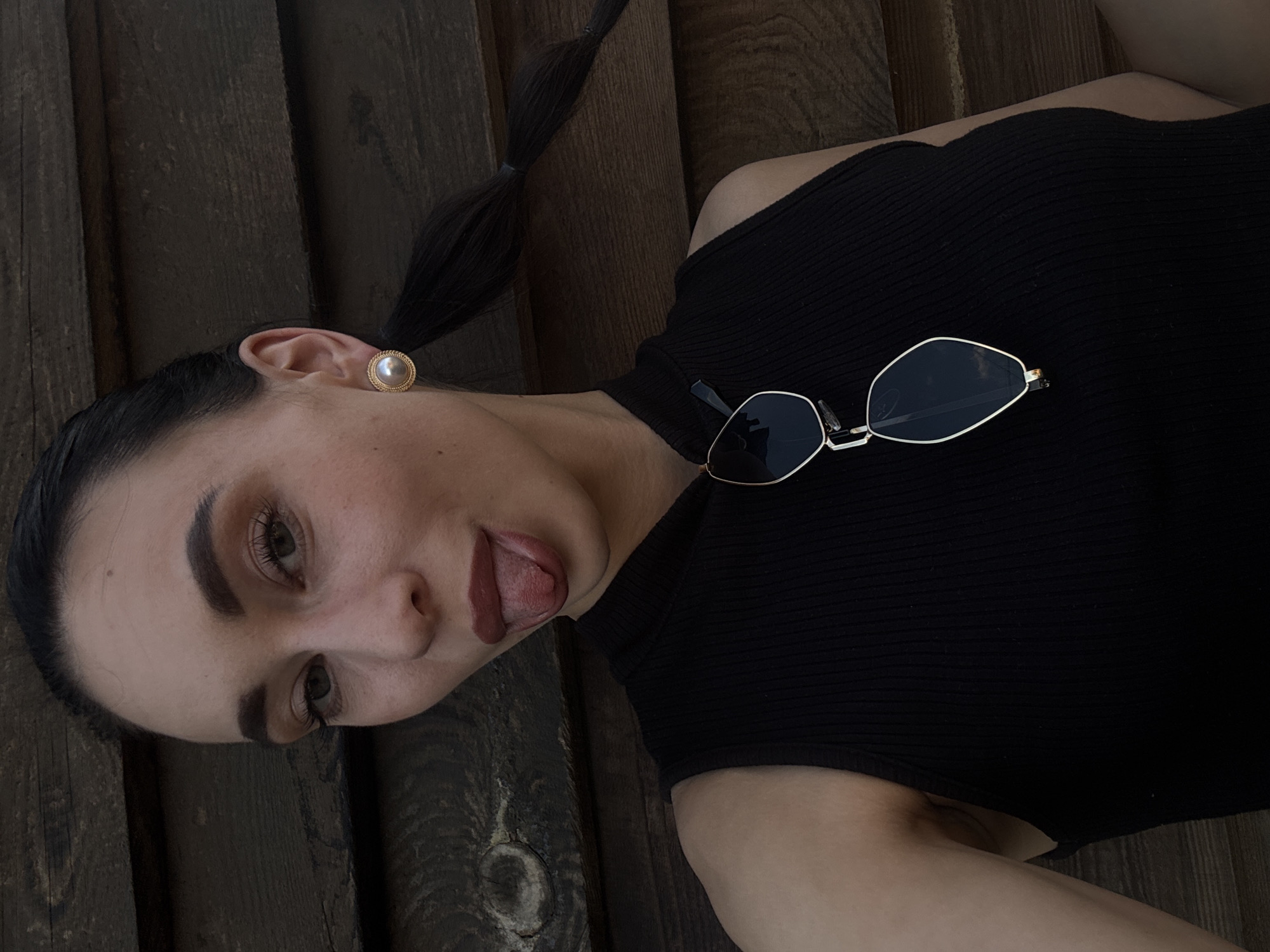 A young woman is lying on a dark wooden surface, likely a deck or floor, with her head tilted towards the camera. She has dark hair pulled back in a ponytail tied with a hair tie that appears like a gold pearl earring. She is wearing a black sleeveless top. A pair of sunglasses with gold frames and black lenses rest on her chest. Her tongue is sticking out playfully. The location is likely outdoors, with the wooden surface suggesting an open space. There is no evidence to pinpoint this image to Chaikovsky, Russia.