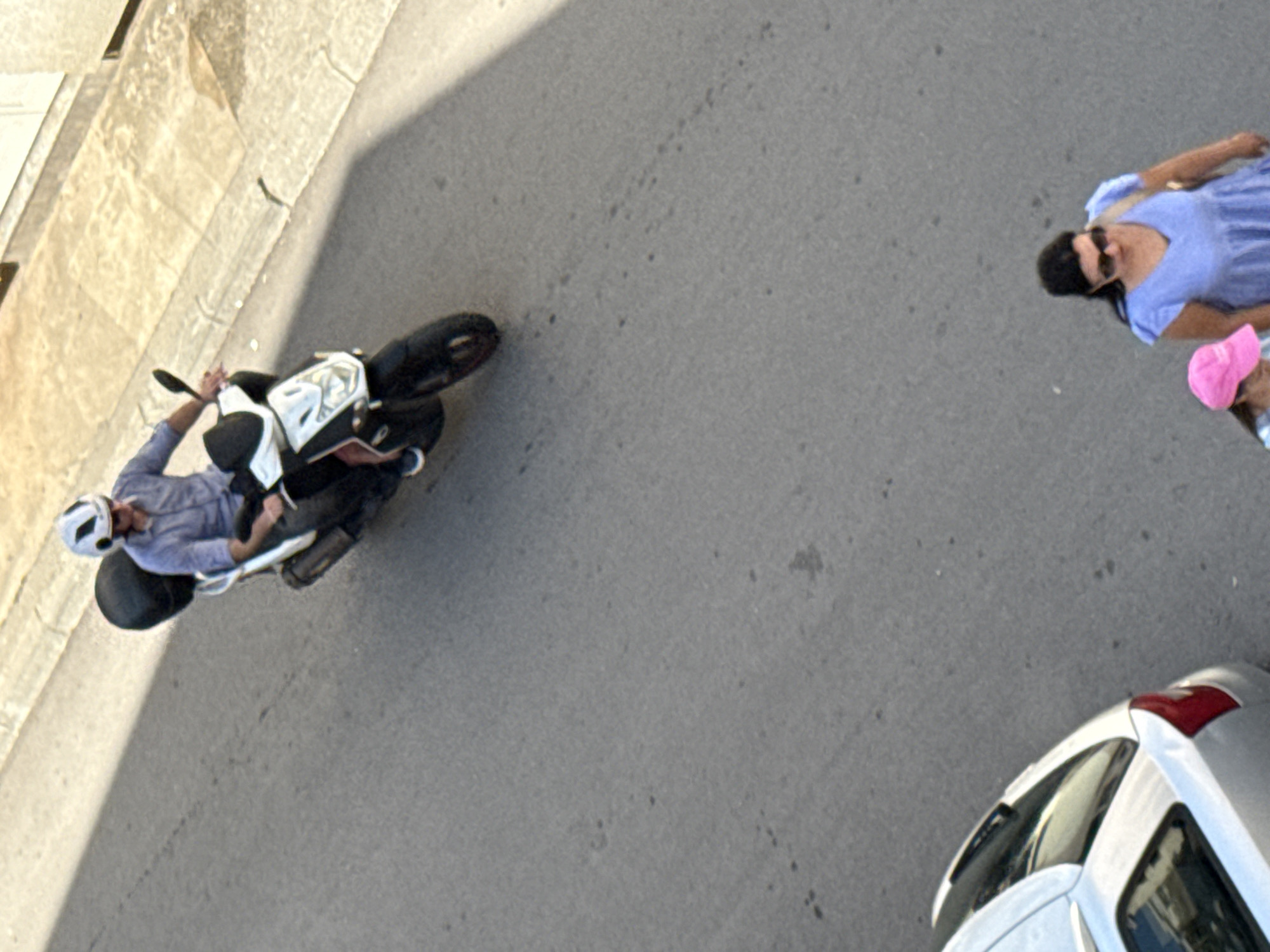 In Budva, Montenegro, a high-angle shot captures a bright, sunny day with a clear sky. The scene unfolds on an asphalt road, with a textured, light-colored concrete wall bordering the left side of the frame. A person, wearing a light purple shirt and a white helmet, is seated on a black and white motorcycle. The motorcycle is positioned on the road, with its kickstand down, suggesting it is stationary.
Further to the right, a woman in a vibrant blue dress and sunglasses walks away from the motorcycle. She is accompanied by a child wearing a pink hat, who is partially visible behind her. The woman appears to be looking back or to the side, though her expression is not discernible. A portion of a silver car is visible in the lower right corner of the image, with its roof and rear spoiler showing. The sunlight casts a distinct shadow of the concrete wall across the asphalt, indicating it is likely late morning or early afternoon. No text is visible in the image.