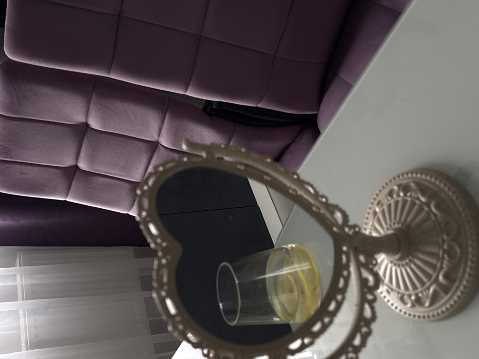 This image depicts an unidentified indoor location. In the foreground, a decorative, heart-shaped vanity mirror, light beige in color with an ornate base, stands on a light-colored table surface. The mirror reflects a clear glass containing a pale yellow liquid with several lemon slices, also resting on the table. In the background, two purple, upholstered chairs with square-tufted backs are partially visible. To the left, a sheer white curtain hangs over a window with a radiator below, adjacent to a darker purple or black curtain. No people are present.