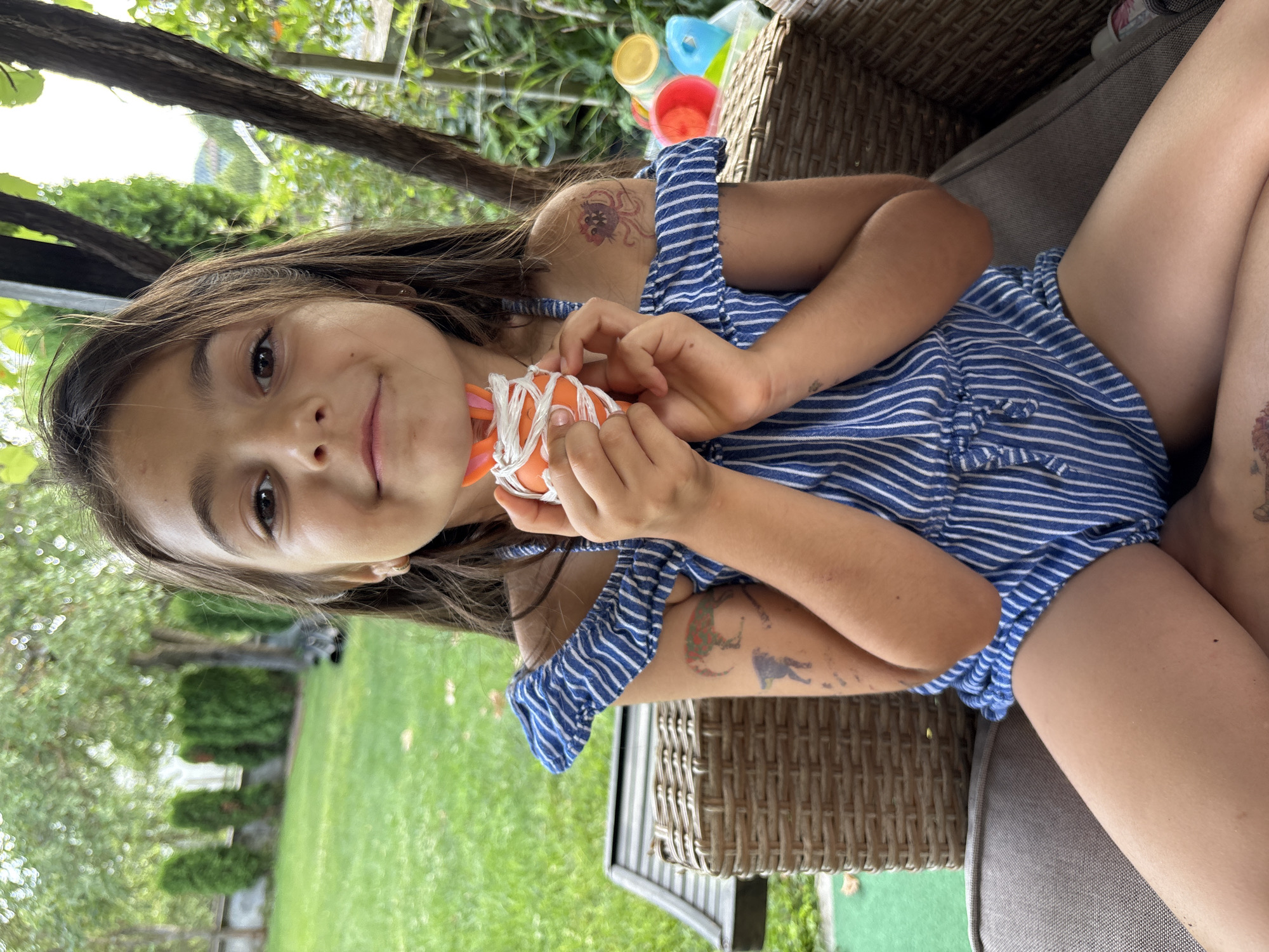 In Oreshak, Bulgaria, a young girl with long brown hair is seated on a wicker chair outdoors. She is wearing a blue and white striped romper. Her left arm is extended towards the camera, holding a small orange object wrapped in white string. She has temporary tattoos on her arms, including a red octopus on her left shoulder and what appears to be a silhouette of an animal on her right arm. She is smiling and looking at the camera. In the background, lush green trees and a lawn are visible, suggesting a sunny day. To the right of the girl, colorful plastic cups are stacked, possibly toys or part of a game. The overall scene conveys a sense of playful outdoor activity.