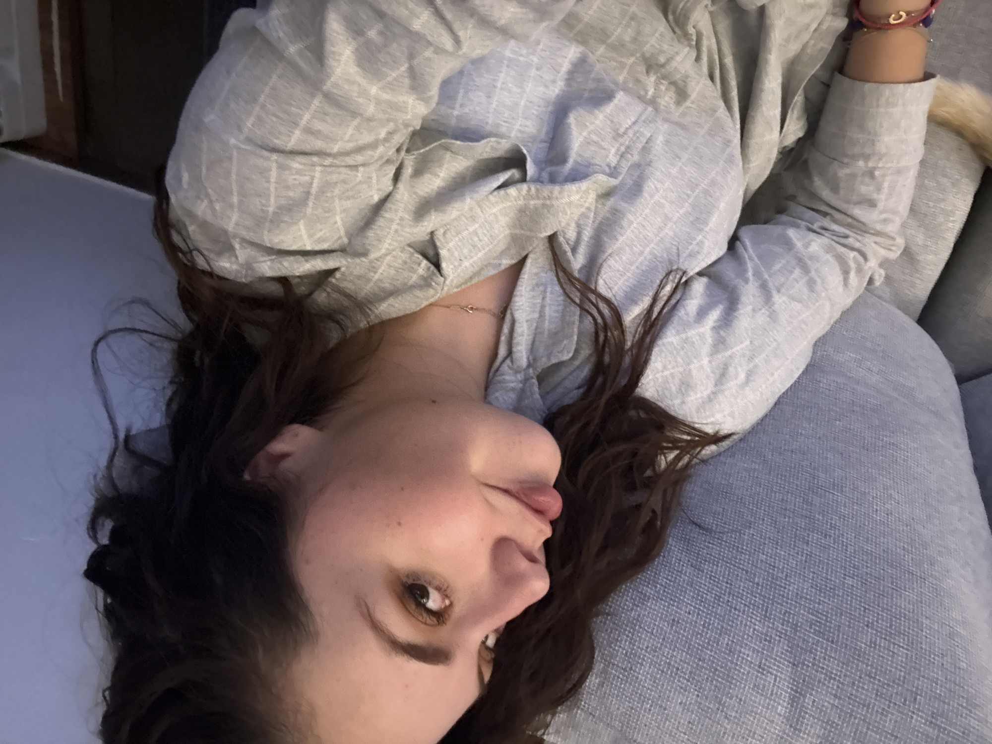 A woman is lying on a light blue sofa. She is wearing a gray and white striped long-sleeved shirt. Her brown hair is spread around her head on the sofa. Her head is tilted slightly, and she is looking at the camera. Her mouth is slightly open, and her tongue is sticking out a little. She is wearing a bracelet on her right wrist. The background is out of focus, but it appears to be a room with a door or window. The location is unknown, but inferred that it is in Mexico City, Mexico.