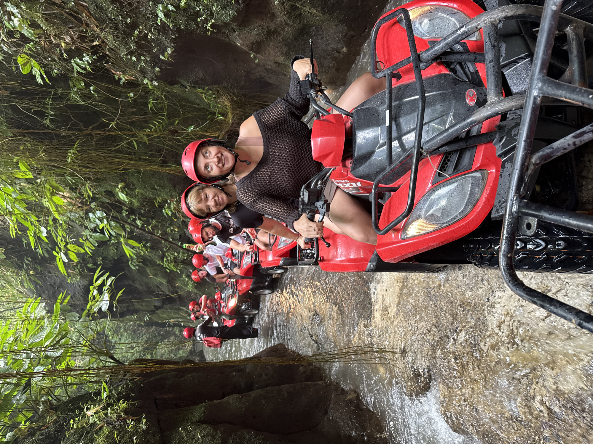 A group of approximately nine individuals wearing red helmets are riding red All-Terrain Vehicles (ATVs) through a shallow, flowing river in a narrow, lush jungle canyon located in Payangan, Indonesia. The immediate foreground features the front of a red KYMCO ATV, with a woman in a black loose-knit top and shorts driving, smiling towards the camera. A second woman, also smiling, is seated behind her as a passenger. Behind them, a line of other red ATVs proceeds through the river, occupied by additional riders and passengers. One individual further back, near the left canyon wall, is standing in the river next to an ATV, wearing black clothing and a black backpack, possibly a guide. The river water is turbulent and light brown, reflecting some ambient light, with splashes around the ATV wheels. Both sides of the narrow passage are defined by steep, dark rock walls heavily covered in dense green tropical foliage, moss, and numerous long, thin hanging vines descending from above. The overall scene depicts an organized recreational activity within a natural, enclosed environment.