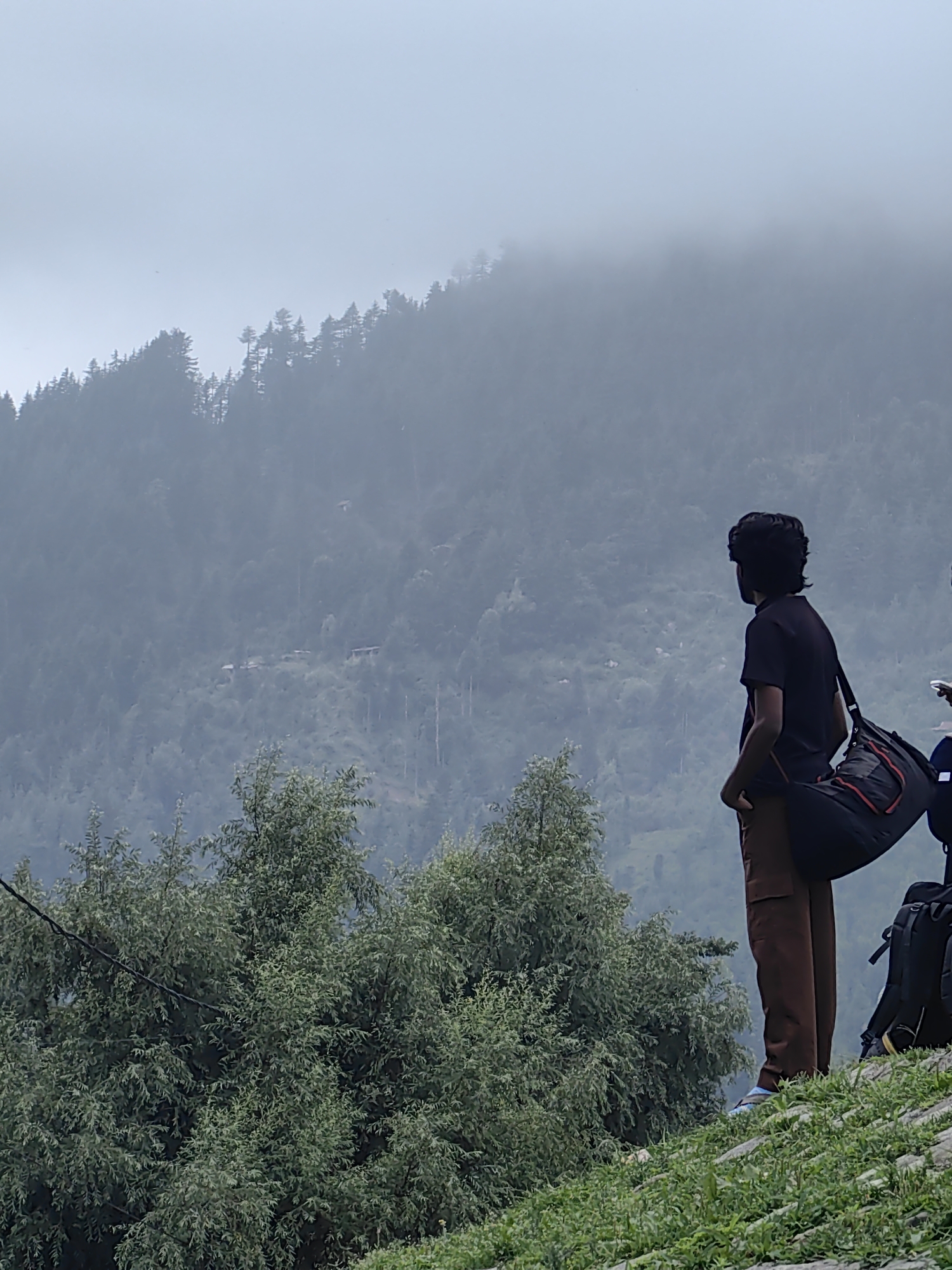 The image captures a man standing on a grassy incline, looking out at a misty, forested mountain landscape. The man is wearing a black t-shirt, brown trousers, and a black and red duffel bag is slung over his shoulder. Behind him, a large backpack is visible. The mountain slopes are densely covered with trees, with patches of fog creating a hazy atmosphere. The sky is overcast and light grey. The scene appears to be set in a natural, outdoor environment, possibly a hiking trail or viewpoint. The specific location in Vashisht, India, is not directly identifiable from the image, but the mountainous terrain and lush vegetation suggest a Himalayan region.
