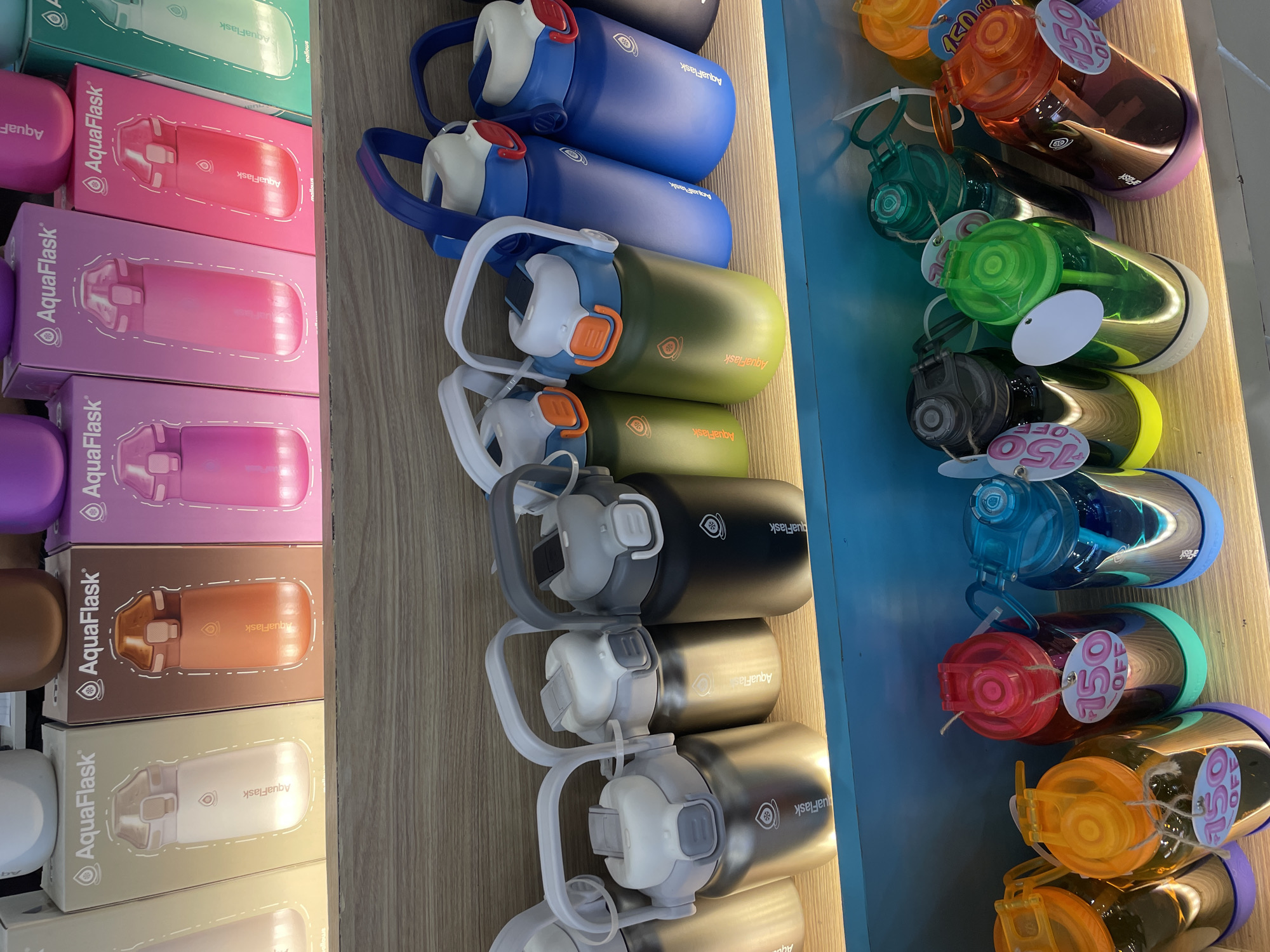 The image displays a retail display of water bottles, likely in a store. The bottles are arranged on two shelves. The top shelf features boxed AquaFlask water bottles in various colors, including cream, brown, pink, and teal. The bottom shelf showcases AquaFlask water bottles without boxes, in colors such as silver, black, green, and blue. Some of these bottles have white or gray handles and lids with orange accents. Further down, a third shelf displays colorful plastic water bottles with prominent "150 OFF" tags. The environment appears to be indoors, with a wooden shelving unit and a blue background on the lower section. There are no people visible in the image. The scene does not provide any specific indicators for a location within Davao City, Philippines.