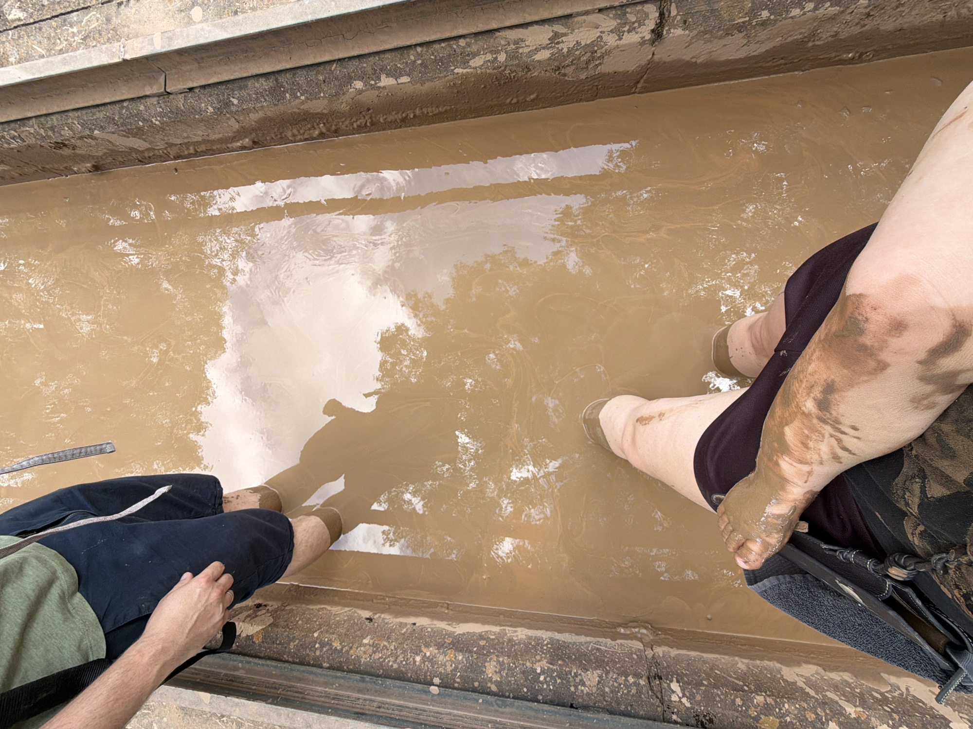 Two individuals are wading through a muddy trench, likely part of the Nahe River flood defense system in Bad Sobernheim, Germany. The trench is filled with murky, brown water and mud, reflecting the surrounding trees and sky. The person in the foreground, viewed from a high angle, is wearing dark shorts and has a muddy leg and foot. The second person, partially visible in the background, is also wading through the mud, their legs submerged up to their knees. The trench walls appear to be made of concrete or stone, with some debris and dirt accumulated on the edges. The overall scene depicts an outdoor environment affected by recent rainfall or flooding, with evidence of human activity in the muddy water.