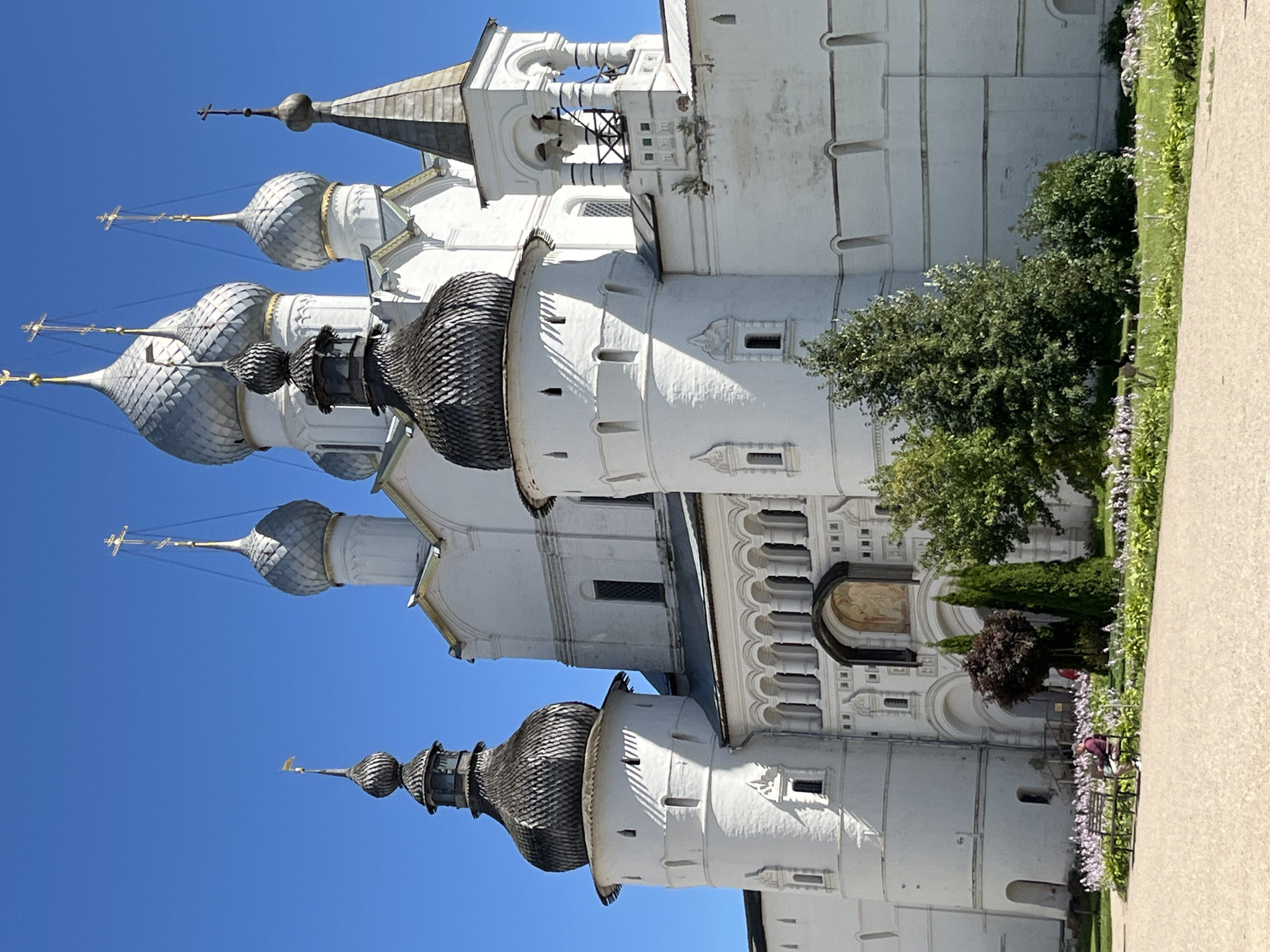 This is a full, eye-level, outdoor, medium shot of the Rostov Kremlin in Rostov, Yaroslavl Oblast, Russia. The Kremlin is a large, white, stone fortress with a bright blue sky overhead. Several onion domes, some of which are silver and some of which are gray, rise above the main structure, topped with golden crosses. The building is detailed with many arches, windows, and decorative elements. In the foreground, a paved walkway curves around a patch of green grass dotted with small pink and white flowers. A dark wooden bench sits on the grass, with a single person wearing a maroon shirt and dark pants sitting on it, facing away from the camera. Several trees, some with dark green foliage and others with lighter green leaves, are planted in front of the Kremlin. The overall scene depicts a sunny day in a historic religious site.