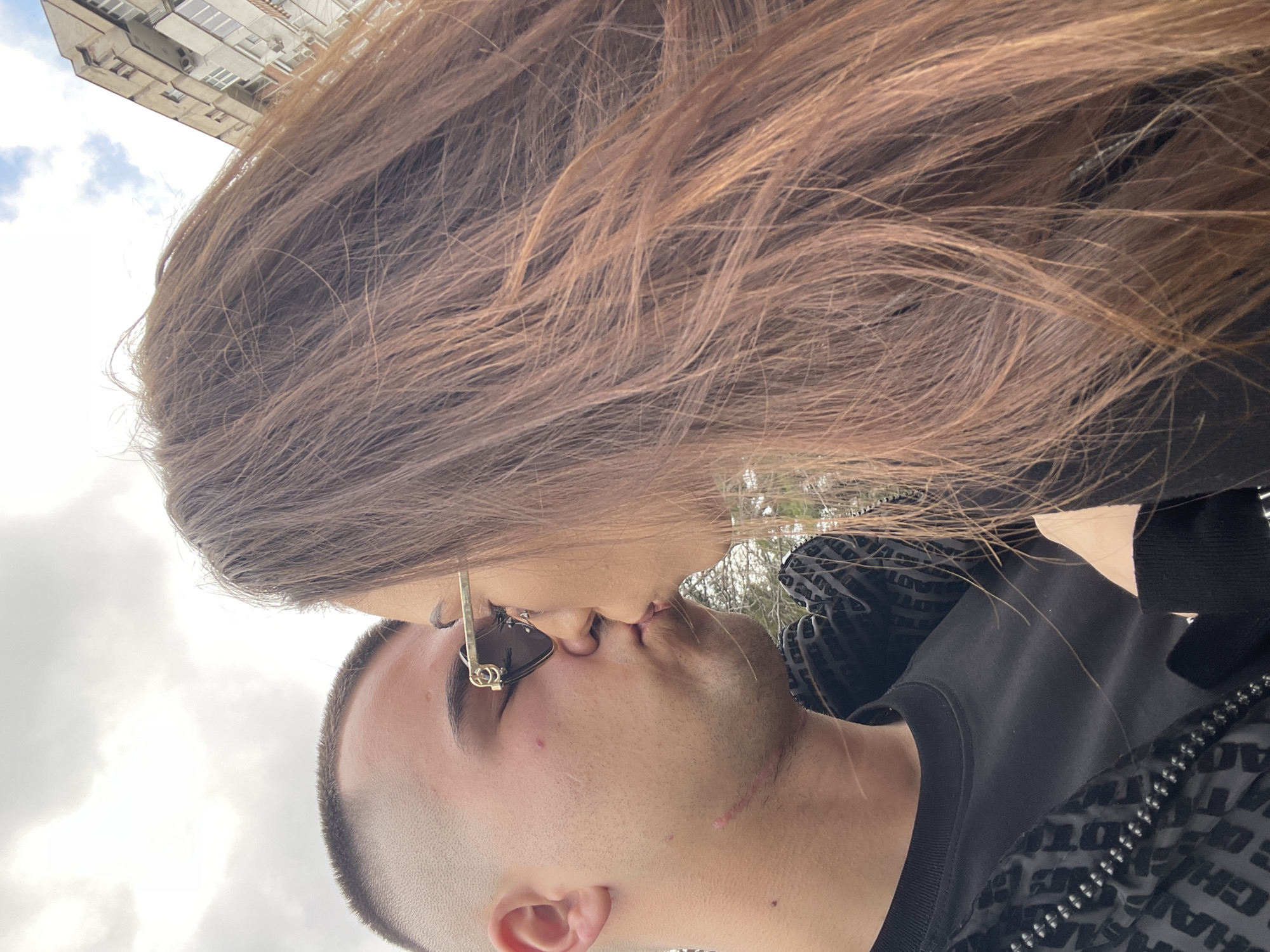 A close-up shot depicts a man and a woman kissing. The man has short, dark hair and is wearing a black jacket with a patterned design and what looks like a plain black shirt underneath. The woman has long, brown hair and is wearing a black top. She also has a gold earring and sunglasses resting on her face, visible just above her nose. The background includes a cloudy sky and a building. Specific location in Veliko Tarnovo, Bulgaria, cannot be determined from the image.