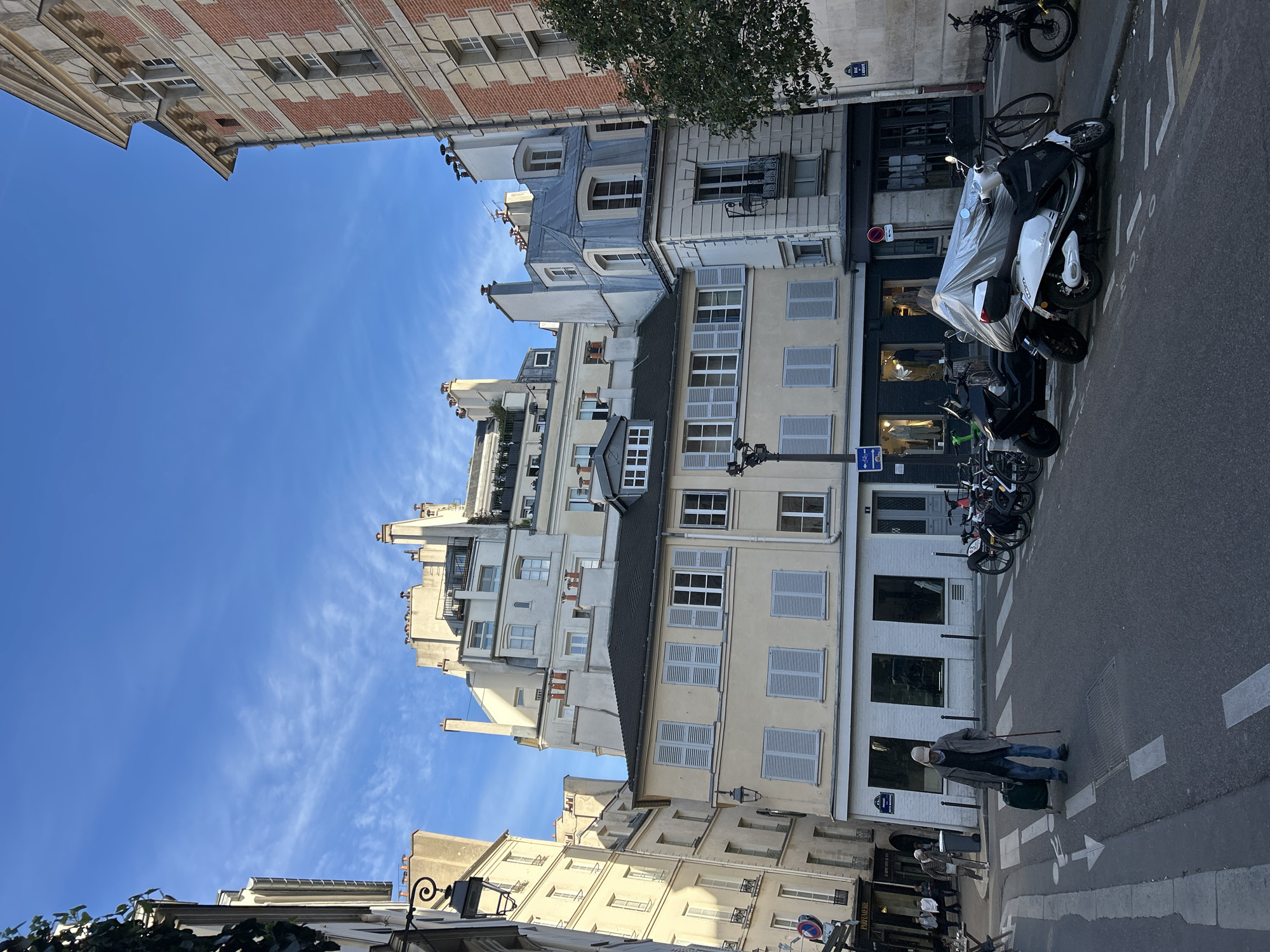 The image depicts a street scene in Paris, France, under a clear blue sky with faint cirrus clouds. Architectural styles indicate a historical European urban setting.
In the foreground, a crosswalk is visible on the asphalt street. A person, dressed in a light-colored cap, dark jacket, and blue jeans, is crossing the street. They are using a cane and pulling a small, dark-colored wheeled bag.
Parked along the side of the street are several motorcycles and bicycles. The buildings lining the street are multi-story, with varying heights and styles. Some have light-colored facades with grey shutters, while others are brick. There are shops on the ground level, with their windows and entrances visible. A street sign is affixed to one of the buildings.