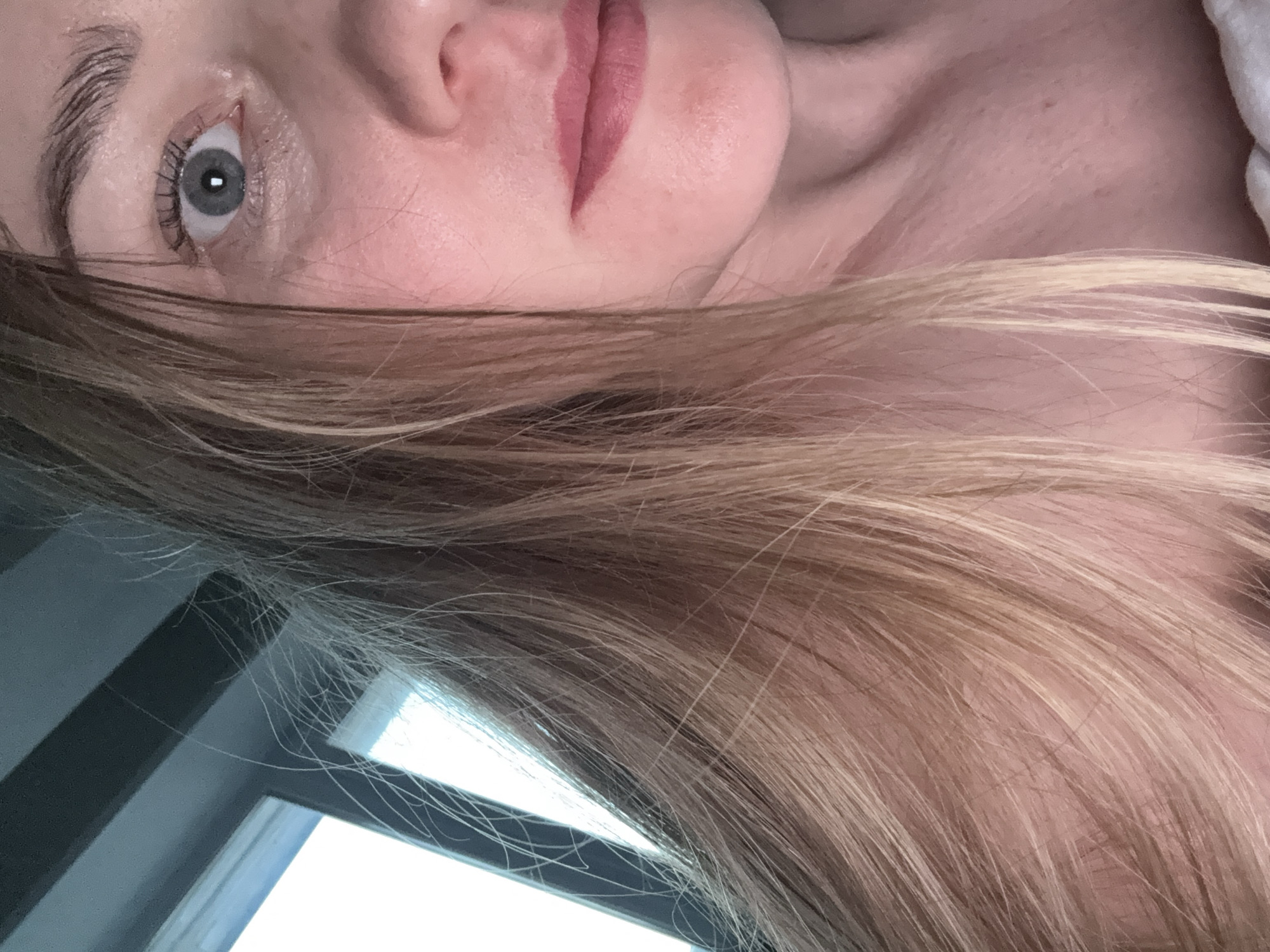 A close-up, upward-angled portrait of a person with fair skin, light brown hair, and light gray-blue eyes. The person's face is partially obscured by their hair. Visible facial features include the eye, nose, and mouth, which is slightly open, revealing pink lips. Part of a window with dark frames and bright light filtering through is visible in the lower left corner of the image. There is no indication of the location being in Chaikovsky, Russia, or any other specific location.
