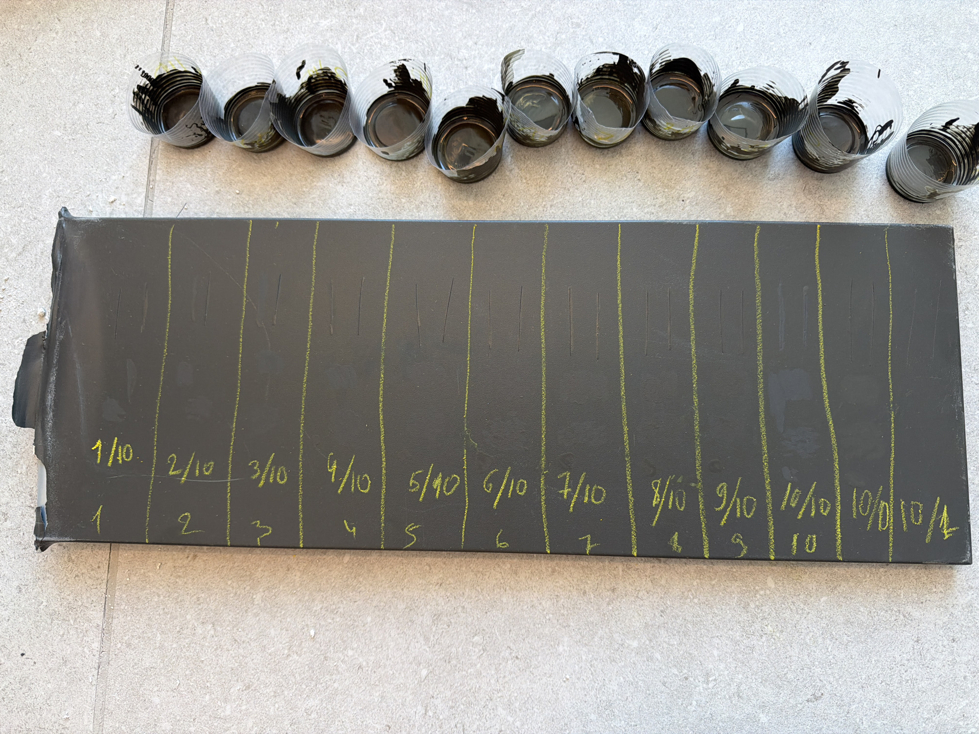 This is an overhead, close-up, eye-level, full shot of a dark gray, rectangular board or panel lying on a light gray, textured, tiled surface. The board is positioned vertically in the frame and has yellow chalk markings on its surface. These markings include numbers written along the left edge of the board, starting from "1" at the top and going down to what appears to be "14" at the bottom. Next to each number, there is a fraction, such as "1/10", "2/10", "3/10", and so on, up to "10/10", and then "11/10", "12/10", "13/10", and "14/10". There are also horizontal yellow lines drawn across the board at intervals, corresponding to the numbered markings.
To the right of the board, there is a vertical line of empty, clear plastic cups, stacked one above the other in a slightly staggered arrangement. The cups are mostly empty, but some of them contain black liquid or residue at the bottom, and a small amount of yellow residue is visible in some cups.
The tiled surface appears to be outdoors, possibly a patio or walkway. The lighting is bright, creating subtle shadows. The scene is devoid of any people or animals. The overall impression is of an experiment or measurement being conducted, perhaps involving the contents of the cups and their relation to the markings on the board. The location of Jerusalem, Israel, is not discernible from the visual information in the image.