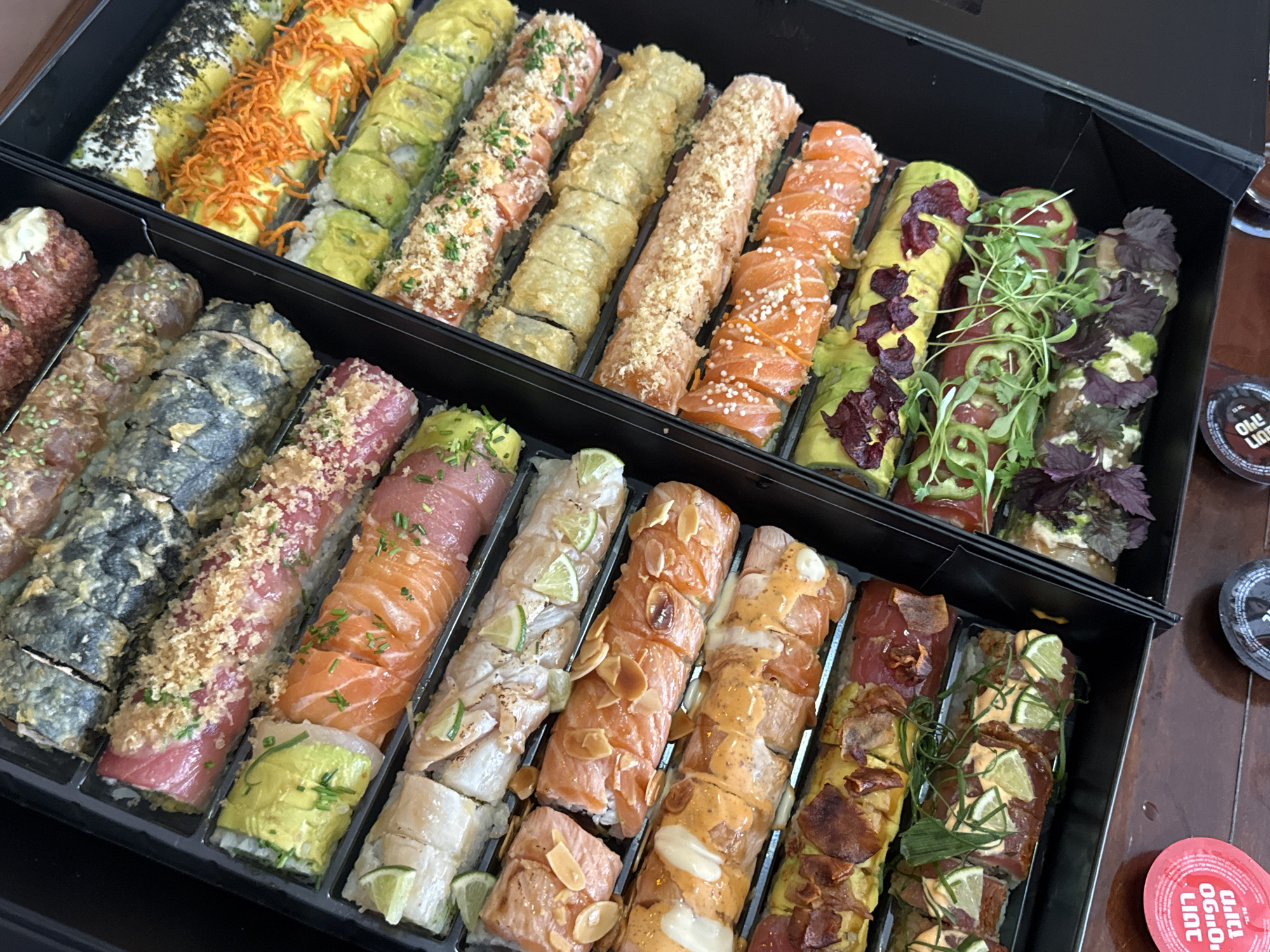 This image shows a close-up, overhead view of two black sushi trays filled with various sushi rolls. The trays are positioned on a wooden surface. The sushi rolls are arranged in rows, with different types of ingredients and toppings visible. Some rolls are topped with avocado, salmon, tuna, and what appear to be crispy fried elements. There are also some red circular containers and dark circular containers on the wooden surface below the trays. One of the red containers has text in Hebrew. The overall scene suggests a food presentation, likely in a restaurant or for a takeaway order. The text on the red container, "רוטב סויה", translates to "Soy sauce", indicating the presence of dipping sauce. The location mentioned, Hof HaCarmel Regional Council, Israel, places this scene within that geographical context.