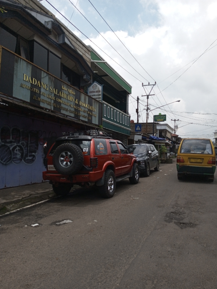 The image depicts a street scene in Cireunghas, Cianjur, West Java, Indonesia, under an overcast sky. A red four-door SUV, possibly a Chevrolet Blazer, is parked curbside on the left, facing away from the viewer. It features a rear-mounted spare tire, a roof rack, an aftermarket rear bumper, and chrome wheels. A "01" decal and an additional graphic are present on its rear window. Immediately behind the red SUV, a dark gray SUV is also parked. Further down the street on the right, a yellow and green minivan is visible. The asphalt road surface shows wear, including cracks and minor potholes.
To the left, a multi-story building features a lower facade with purple-blue paint and graffiti. A prominent sign on the upper floor reads "Law Office DADANG SALAHUDIN & ASSOCIATES ADVOCATES AND LEGAL CONSULTANT" and includes the address "JL. MARIWAH NO. 54 CIREUNGHAS - CIANJUR". The building has dark-tinted windows on the upper level and a green-colored roofline or facade accent. Numerous utility wires crisscross overhead, supported by multiple utility poles. A blue triangular road sign and a green sign with white text are visible further down the street on the right. No individuals are present in the scene.