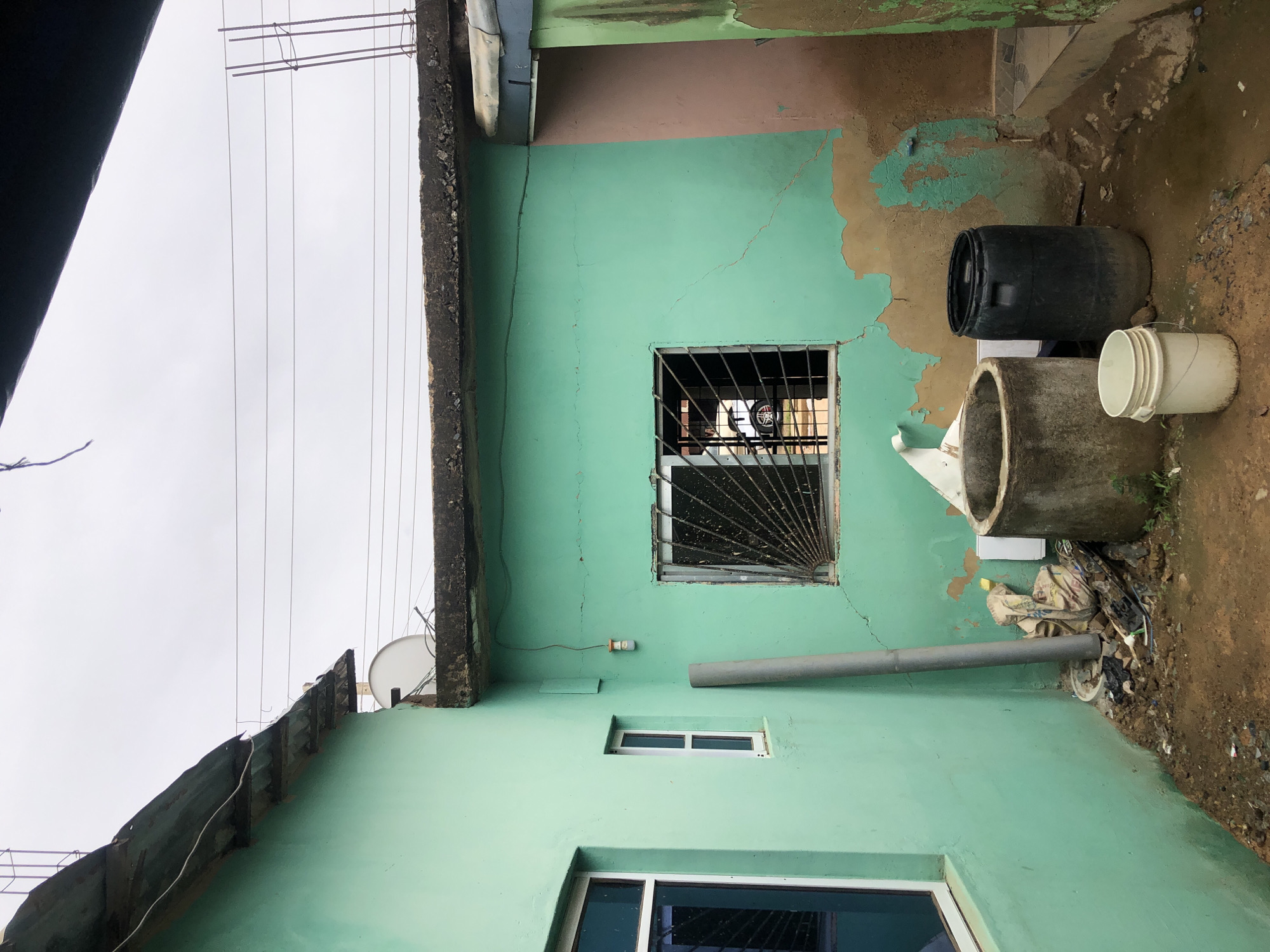 The image captures the exterior side of a building, primarily painted in a faded mint green color, seen from a low angle looking upwards. The building shows signs of wear, with patches of peeling green paint revealing a brownish undercoat, and several fine cracks visible on the wall surface.
Three windows are visible: a larger rectangular window at the bottom with dark frames, a smaller rectangular window with white frames directly above it, and a square window with a black metal security grille further up the wall. Through the security-grilled window, part of a vehicle wheel (possibly a motorcycle or bicycle wheel) and other indistinct objects can be seen inside.
At ground level, next to the building's base, there are various items: a black plastic barrel lying horizontally, a large concrete pipe or culvert standing upright, and a white plastic bucket. The ground itself is uneven, muddy, and covered with scattered debris and trash. A grey vertical downspout pipe runs along the wall, leading to the ground, and another shorter, horizontal grey pipe extends from the wall near the grilled window. A small, simple light fixture is also attached to the wall.
Above the building, the sky is overcast and appears bright white. Multiple thin electrical wires or clotheslines stretch across the sky. The top edge of a concrete structure with exposed rebar is visible, along with a portion of a corrugated metal roof or awning. A small white satellite dish is mounted on an upper section of the building.
The scene depicts a utilitarian and somewhat weathered environment, characteristic of an urban or peri-urban area, likely during daytime under an overcast sky. There are no people visible, and no specific activities are taking place. No visible text is present in the image.