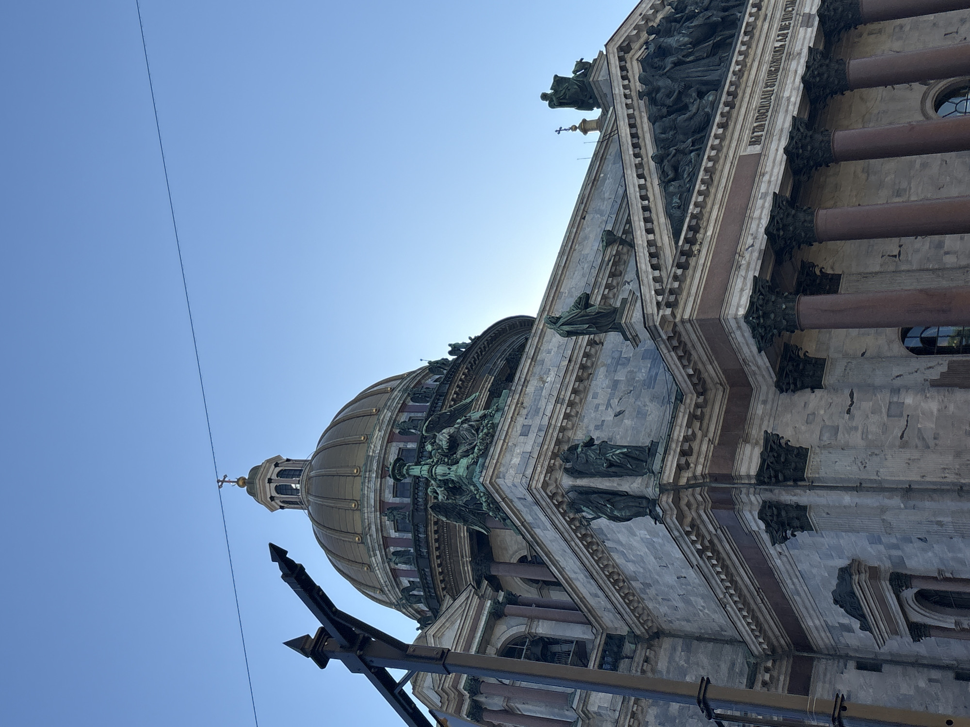 This low-angle, outdoor photograph captures the upper sections of Saint Isaac's Cathedral in Saint Petersburg, Russia, under a clear, bright blue sky. The prominent feature is the large, gilded dome, topped by a smaller cupola with a cross. Below the main dome, multiple tiers of the cathedral are visible, featuring light grey stone facades with dark reddish-brown columns. Numerous bronze or copper statues with green patina, depicting angels and other figures, adorn the various ledges and roofs, including a large sculptural relief within a pediment on the right side of the structure. Below this pediment, dark columns are visible, supporting the upper facade. A horizontal frieze above the columns displays text in Cyrillic characters. In the foreground, a dark metal street lamp structure with pointed finials partially obscures the left side of the cathedral. A thin, dark utility wire spans horizontally across the upper portion of the frame, passing above the dome against the blue sky. No people are actively depicted in the scene, and the environment is characterized solely by the architectural subject and the unclouded sky.
