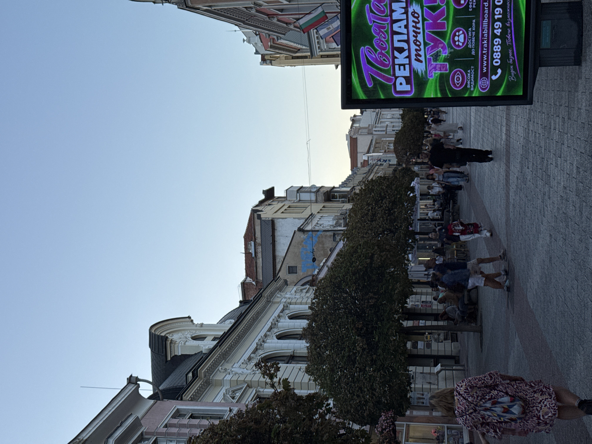 The image shows a street scene in Plovdiv, Bulgaria, likely on a pedestrianized street given the cobblestone pavement and presence of shops and people strolling. The architecture features ornate buildings with classical detailing and mansard roofs, characteristic of older European cities. The sky is a pale blue, indicating either early morning or late afternoon light. A large digital billboard with green and purple lettering dominates the right side of the frame, displaying advertisements in Bulgarian. The text on the billboard includes "Твоята реклама точно тук!" which translates to "Your advertisement right here!". It also shows a website and phone number. People are walking in both directions, some singly and others in small groups. A woman in the foreground on the left is walking away from the camera, wearing a patterned dress and carrying a backpack. Trees line the street, providing shade. Flags, including the Bulgarian flag, are visible on one of the buildings. The overall impression is a lively urban environment.