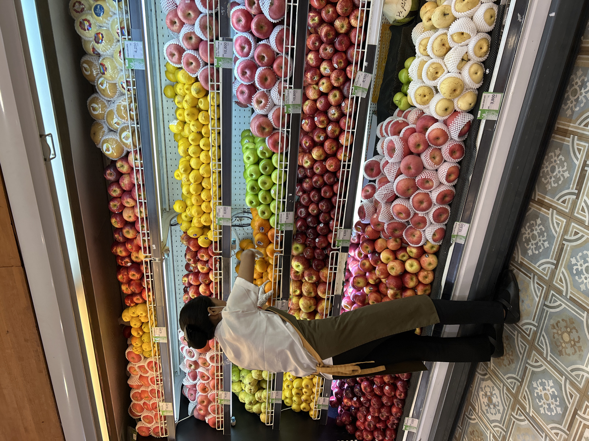 In the produce section of a grocery store in Ubud District, Indonesia, a woman wearing a white shirt, an apron, black pants, and black shoes is seen arranging fruit on a refrigerated display shelf. She has dark hair pulled back in a bun and wears a white face mask. The shelves are filled with a variety of fruits, including red apples, green apples, pears, lemons, and oranges. The floor is tiled with a decorative pattern. The lighting suggests an indoor retail environment.
