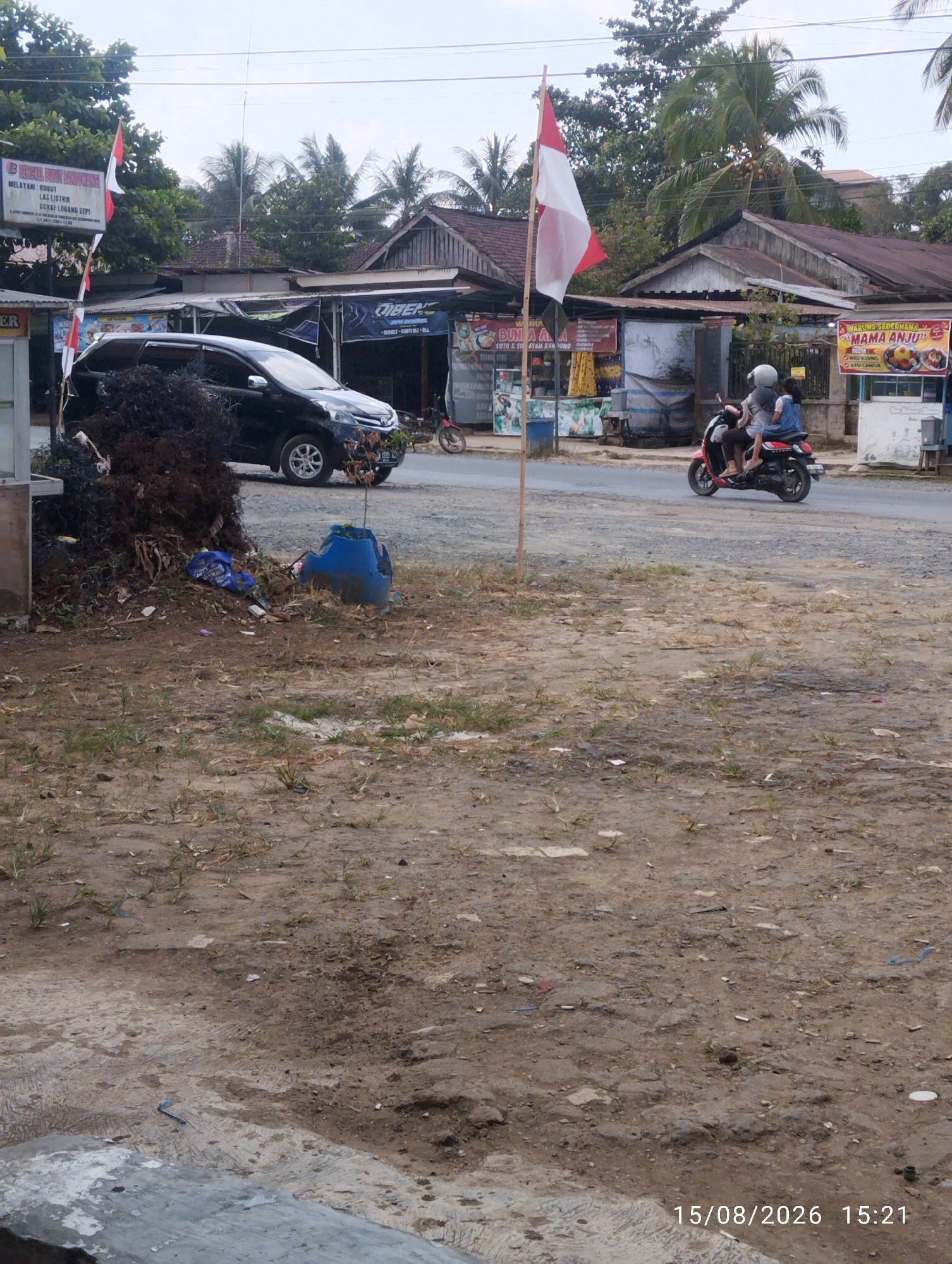 The image depicts a street scene in Tegal Rejo, Indonesia, on August 15, 2026, at 15:21. In the foreground, a dirt and gravel lot is visible with sparse vegetation. A blue plastic container, broken and overturned, lies on the ground near a large pile of debris. In the middle ground, a black minivan is parked on the asphalt road. Further down the road, a red and black scooter carries two individuals, a driver and a passenger, both wearing helmets and casual clothing. They are traveling away from the camera.
To the left of the road, a pole with Indonesian flags is planted, and a sign for "BENGKEL MOBIL KANDANG" which translates to "CAR WORKSHOP KANDANG" is mounted. Next to this workshop, there is a food stall advertising "BUNGA ALA-ALA SOTO & AYAM KAMPUNG" (Flower Ala-Ala Soto & Village Chicken). Across the street, another food stall is visible with a sign that reads "WARUNG SEDERHANA MAMA ANJU" (Simple Stall Mama Anju), also advertising food items like "NASI KUNING" (Yellow Rice) and "KUAH LAMPU" (Lamp Broth). Buildings with tin roofs line the street, with palm trees and other vegetation in the background. The sky is overcast.