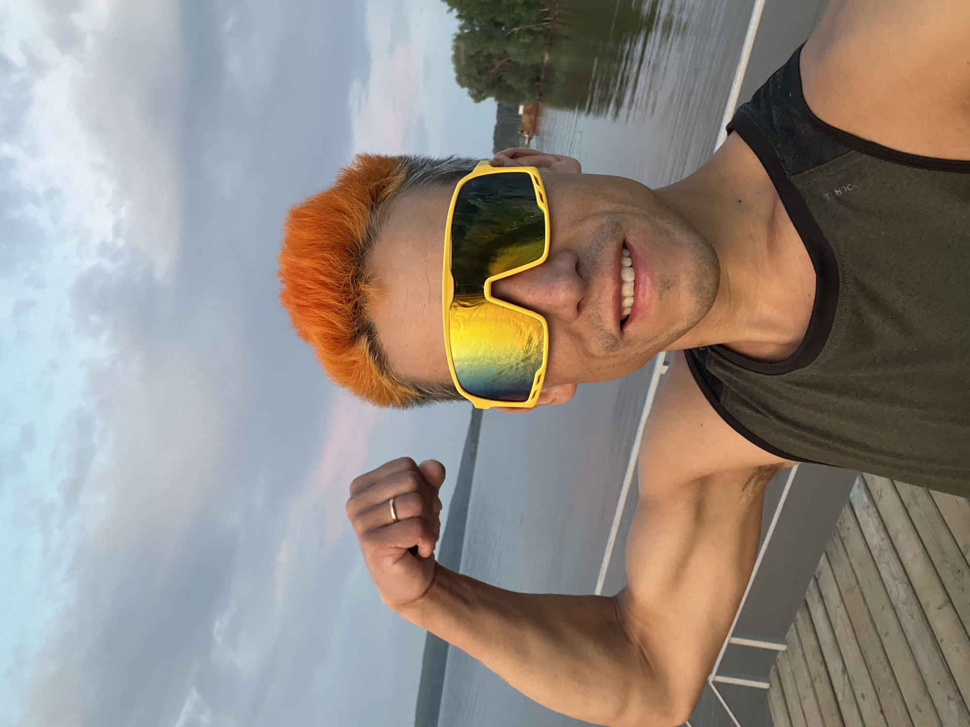 A man with bright orange hair and large yellow mirrored sunglasses is flexing his bicep on a wooden dock. He is wearing a dark green tank top. The background shows a wide body of water with trees and a cloudy sky. This scene is consistent with the waterfront areas of Chaikovsky, Russia, a city located on the Kama River.