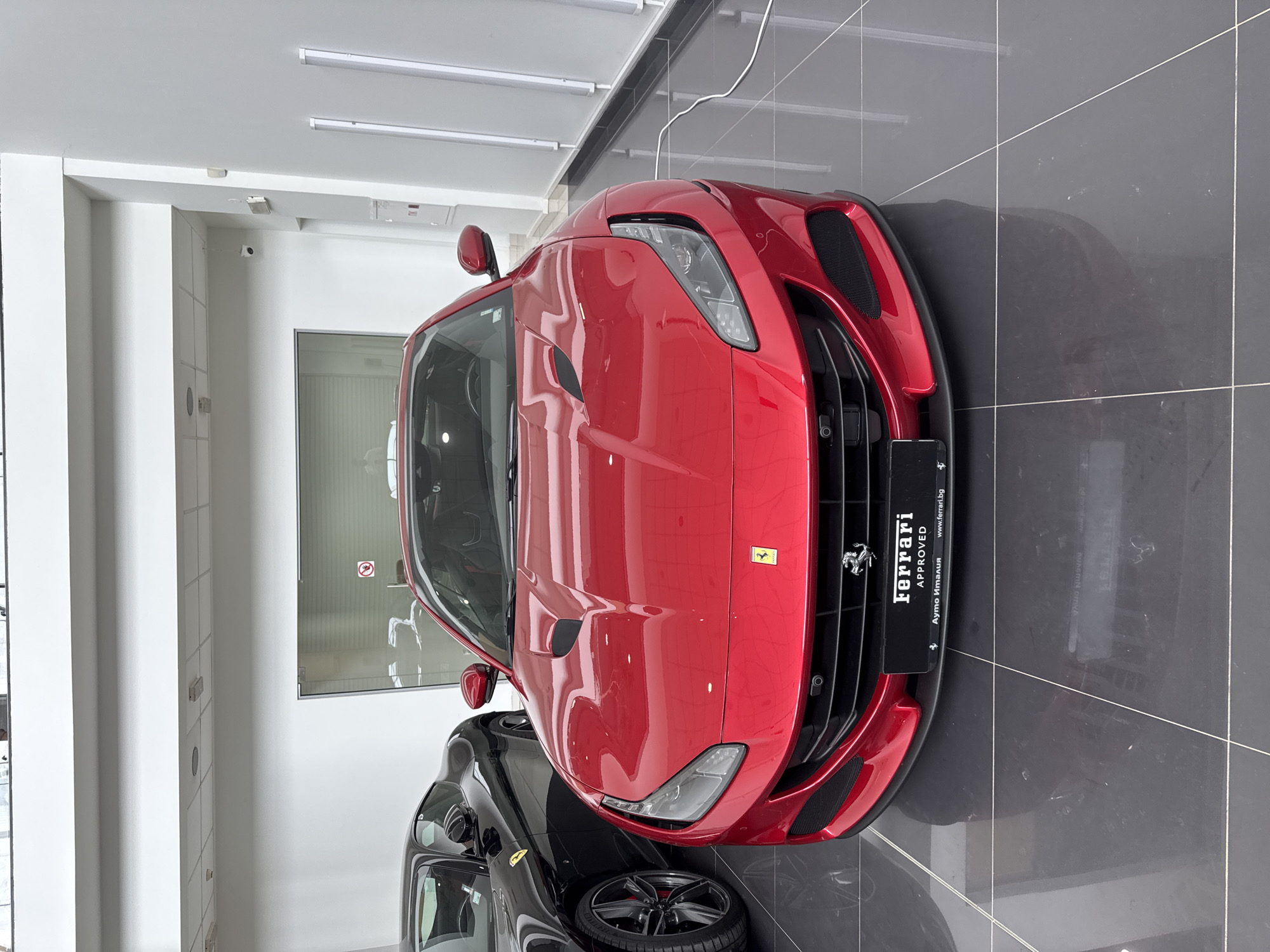 The image captures the interior of a luxury car showroom, specifically a Ferrari dealership. The setting is modern and meticulously clean, featuring pristine white walls, a white ceiling equipped with linear artificial lights, and a dark grey, glossy tiled floor that reflects the vehicles and overhead lighting.
The primary subject is a vibrant red Ferrari sports car, prominently displayed in the foreground. Its sleek, aerodynamic lines and lustrous paint finish are accentuated by the bright, even illumination. Partially visible at the bottom of the frame is the front section of a second, black sports car, likely also a Ferrari, indicating a display of multiple high-end vehicles. There are no people visible in the scene.
The cars are presented for display, consistent with a dealership environment. Artificial lighting ensures consistent brightness. A small security camera is mounted on a white column in the background.
Visible text on a display plate attached to the front of the red Ferrari includes "FERRARI," "APPROVED," "Ауто Италия" (Auto Italy), and the website "www.ferrari.bg." The iconic Ferrari 'prancing horse' emblem is visible on this plate and the car's grille, while a yellow Ferrari shield emblem adorns the front fender. A faint "no smoking" symbol is reflected in a glass partition in the background. The ".bg" domain and "Ауто Италия" strongly indicate the dealership's location in Bulgaria, aligning with the provided context of Sofia. The overall atmosphere conveys luxury, exclusivity, and automotive prestige.