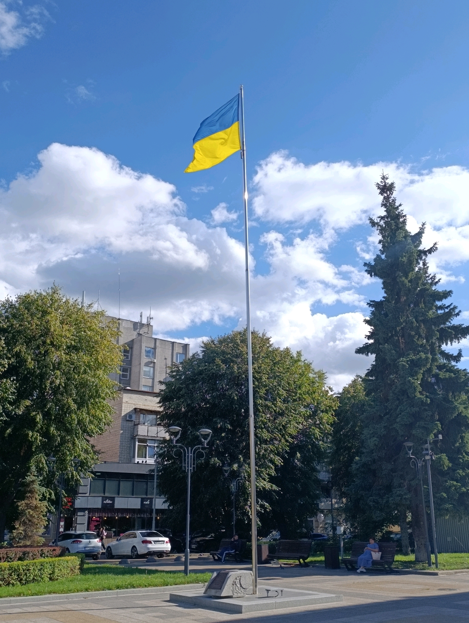 The image captures a view of a city square in Cherkasy, Ukraine, on a bright, partly cloudy day. Dominating the foreground is a tall flagpole flying the Ukrainian flag, its blue and yellow colors vibrant against the sky. The flagpole stands on a concrete pedestal with a plaque.
To the left of the flagpole, several deciduous trees with green foliage line a sidewalk. Behind the trees and flagpole, a multi-story building with a gray facade and numerous windows is visible, its rooftop adorned with antenna-like structures.
To the right of the flagpole, a tall, dark green coniferous tree, likely a spruce or fir, stands prominently. Further to the right, another street lamp and more greenery suggest a park-like setting.
In the midground, several parked cars, including a white sedan and a silver SUV, are visible on a street or parking area. People are present in the scene: a few individuals are seated on park benches, and others appear to be walking or standing near the parked cars and a storefront. The storefront, with large windows, has signage that includes the word "Cafe" and possibly other Ukrainian text. The overall environment suggests a public urban space with a combination of architectural elements and natural greenery.