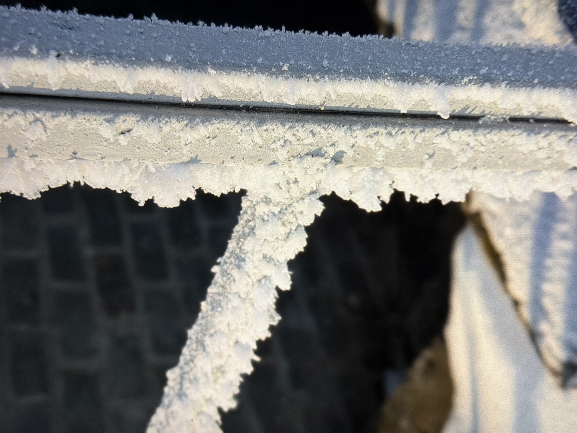 The image shows a close-up of frost crystals covering a metal railing. The railing appears to be outdoors, and in the background, a blurred brick wall is visible. The lighting suggests it is daytime, with sunlight illuminating the frost and casting shadows. The environment is cold, as indicated by the presence of frost. There are no people or animals visible in the image. The scene is one of natural winter conditions.