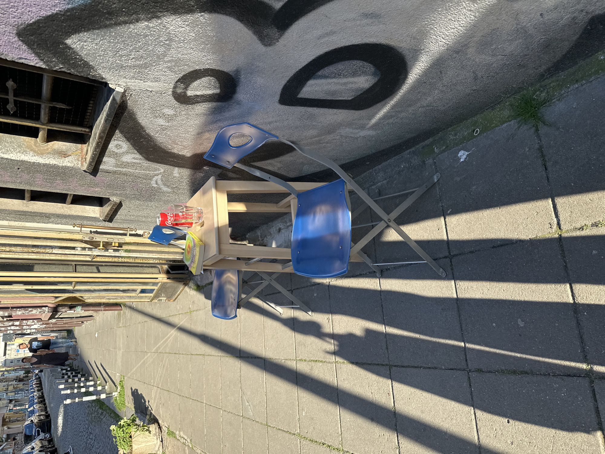 The image depicts an outdoor seating area on a sidewalk in Sofia, Bulgaria, with a building adorned with graffiti to the right. Two blue folding chairs are placed around a small wooden table, on which a book, a Coca-Cola can, and a glass of water are visible. The scene is illuminated by strong sunlight, casting long shadows from the chairs and table across the concrete pavement. In the background, a street lined with parked cars and a few people walking on the sidewalk is visible.