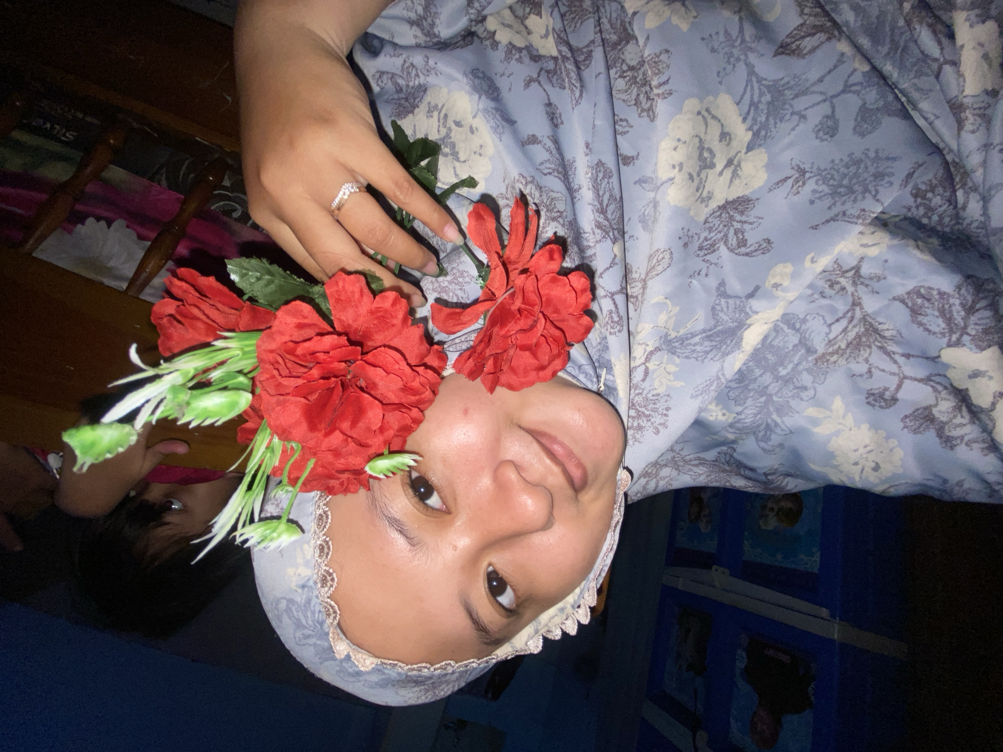 A medium shot depicts a person with brown skin, wearing a headscarf with a floral print and lace trim. Red artificial flowers adorn the left side of the person's head. A ring is visible on the person's left hand. The person is smiling slightly. To the left, the face of a child is partially visible. The background includes furniture and a blue structure with cartoon images, and the overall lighting is dim. The image location might be Gelam, Indonesia.