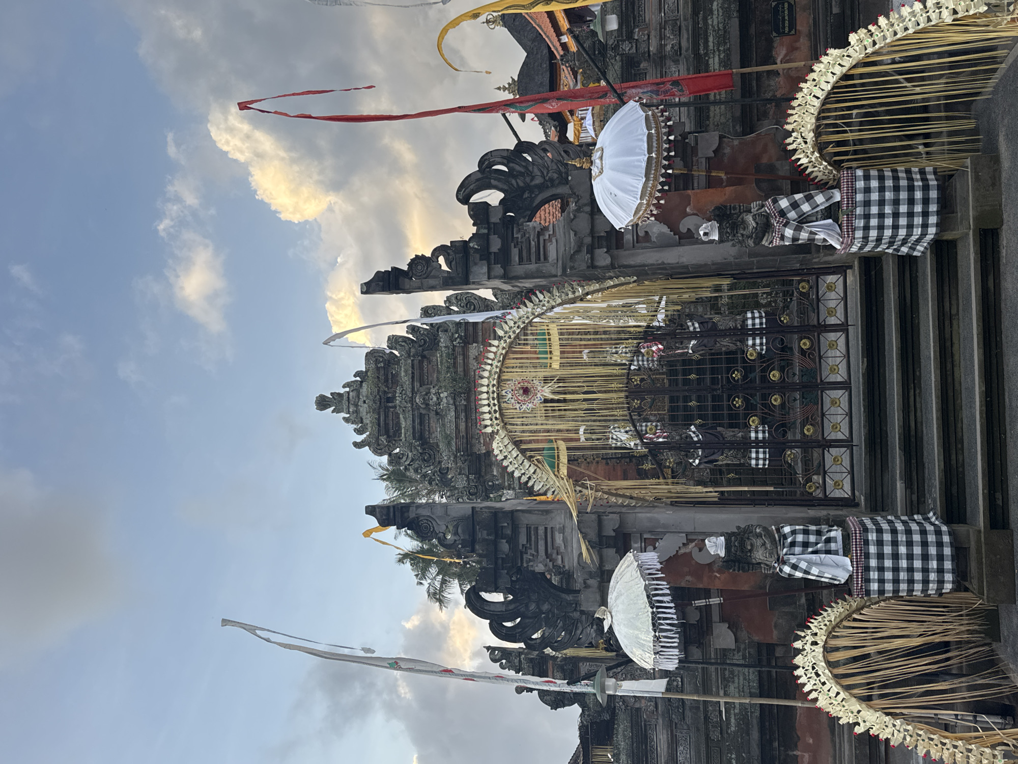 A frontal, medium shot captures the ornate entrance to a temple in Blahbatuh, Indonesia, under a partially cloudy sky. The temple gate is a dark, weathered stone structure with intricate carvings and multiple tiers. A large, arched gateway is adorned with golden, thread-like decorations and bamboo-like strands. Flanking the entrance are two stone statues, each dressed in traditional black and white checkered sarongs and white headdresses. Above each statue, a white, fringed umbrella is positioned.
Decorative flags are visible throughout the scene: a tall white flag with red and green accents stands to the left, a vibrant red flag with white accents is positioned to the right of the central gate, and a yellow flag is seen further back. Golden, fan-shaped decorative elements, woven from natural materials, frame the base of the entrance on either side. The ground in front of the gate consists of stone steps. Beyond the main gate, a glimpse of the temple's interior reveals more architectural details and the muted colors of other structures. The overall atmosphere is serene and cultural, indicative of a religious or ceremonial site.